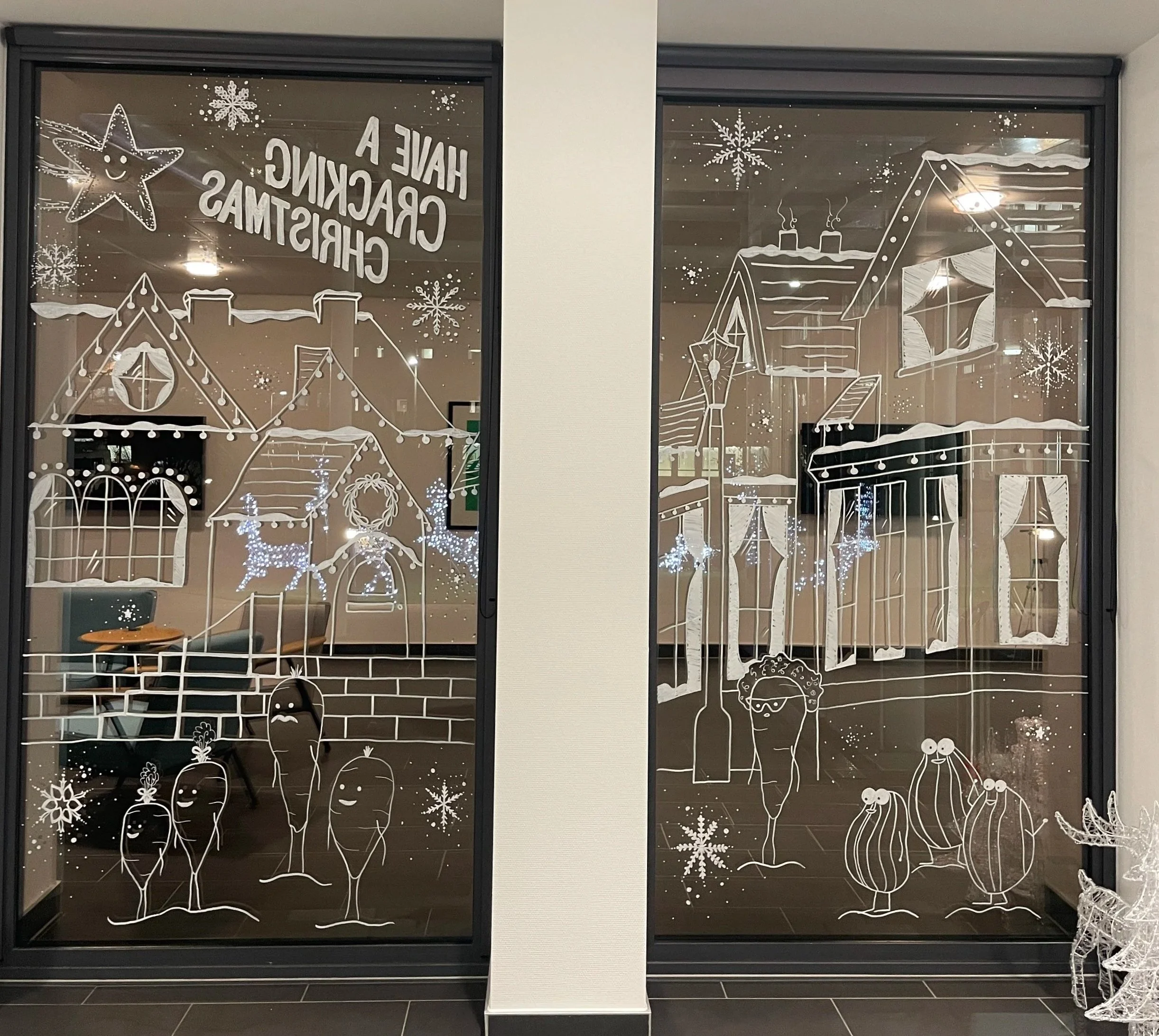 Decorative holiday window art with white line drawings of houses, trees, street lamps, snowflakes, and cartoonish vegetables with facial expressions, including carrots and onions, along with the phrase 'Have a cracking Christmas' on the left window.