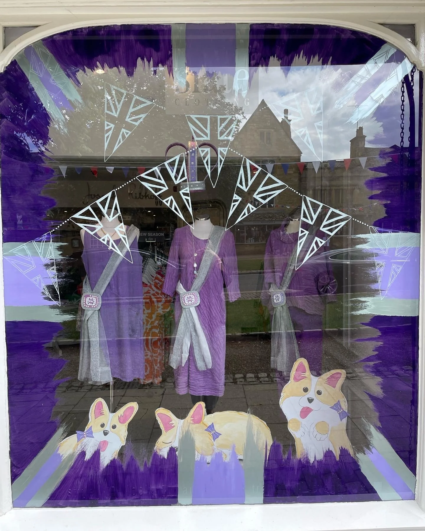 Store window display featuring purple dresses, white and purple scarves, and paper cutouts of playful corgi dogs with purple bows, decorated with white star-shaped banners and purple borders.
