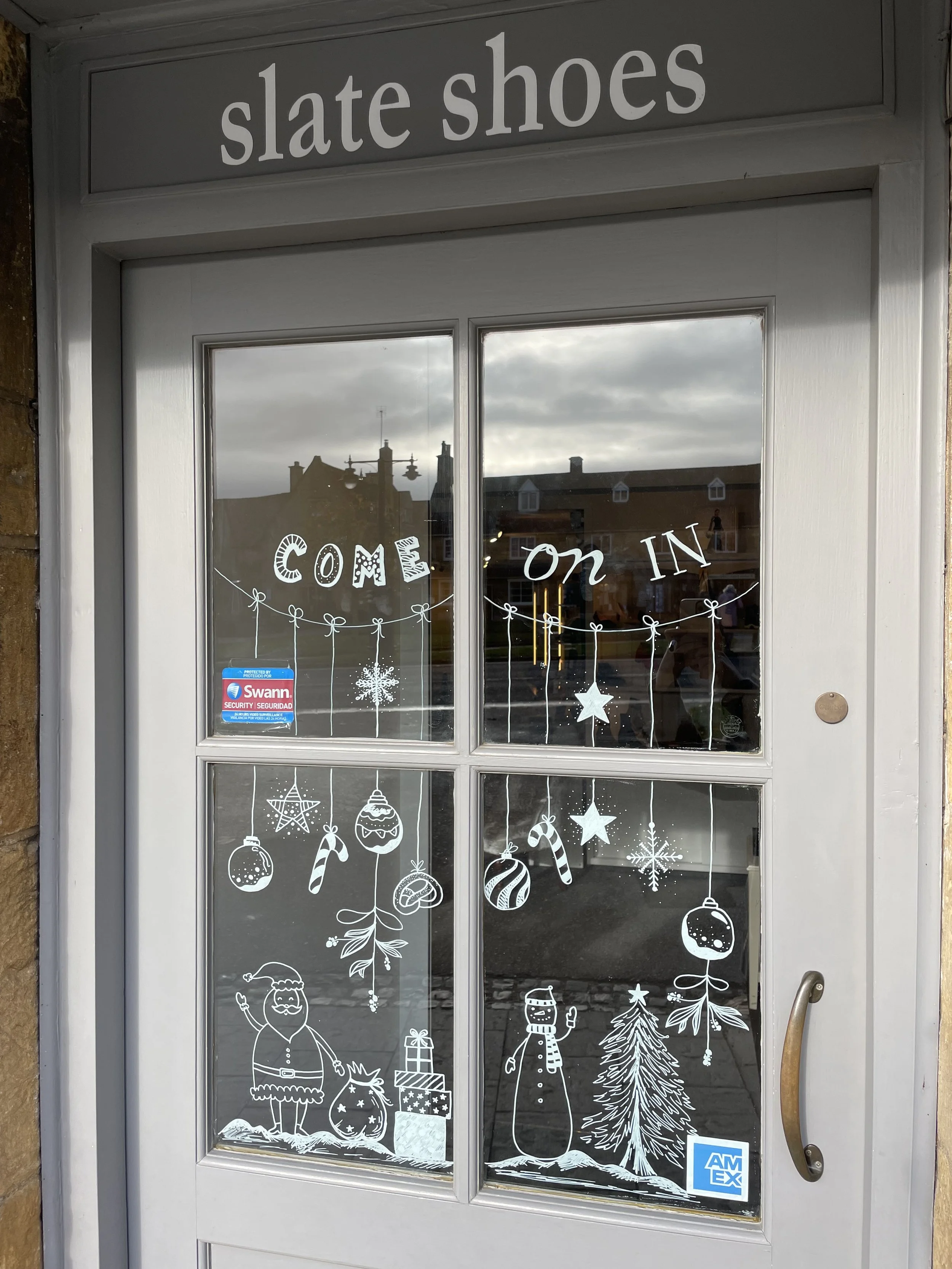 Decorative storefront window with white holiday-themed illustrations including Santa, a snowman, Christmas ornaments, stars, snowflakes, a Christmas tree, and gifts. The text 'Come on in' is displayed across the window. The shop is named 'slate shoes