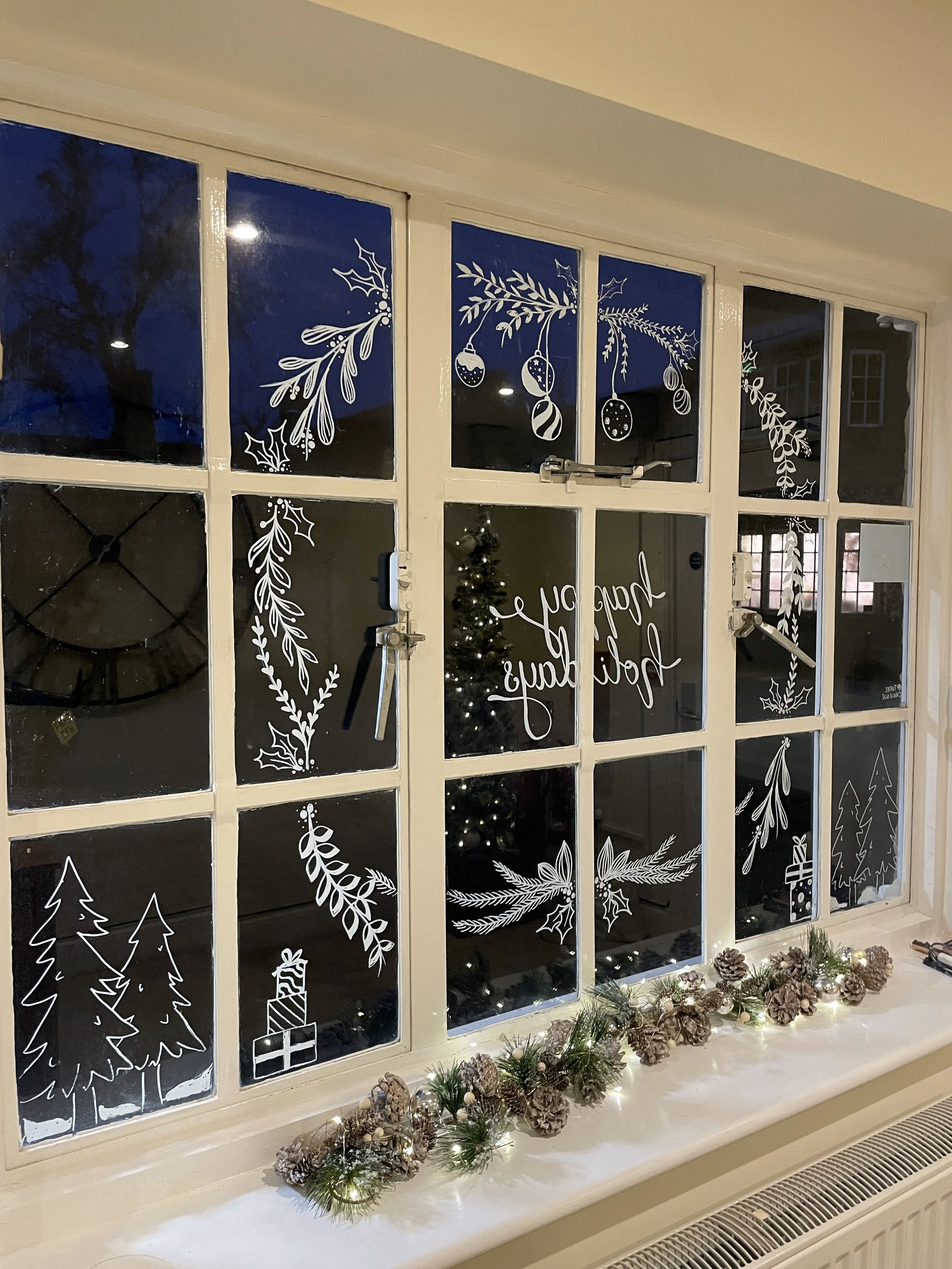 Decorated window with white holiday-themed drawings including pine branches, Christmas balls, holly, pine trees, a gift, and berries. A decorated Christmas tree is visible inside, and a festive garland with pine cones, lights, and greenery is on the 