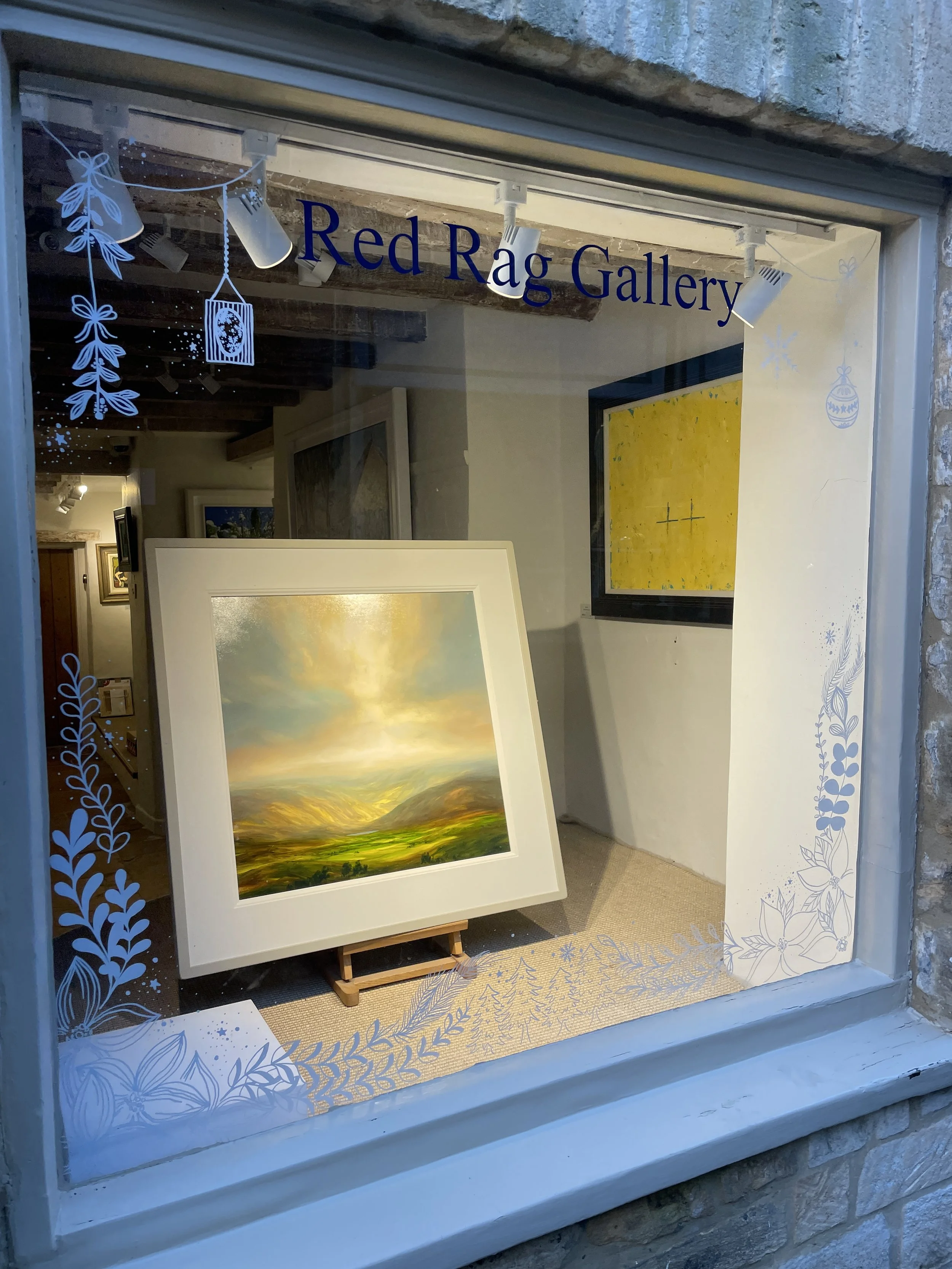 View of an art gallery window with the sign 'Red Rag Gallery' at the top. Inside, there are paintings on display, including a landscape with mountains and a cloudy sky, and another yellow abstract piece. Decorations with winter-themed blue and white 