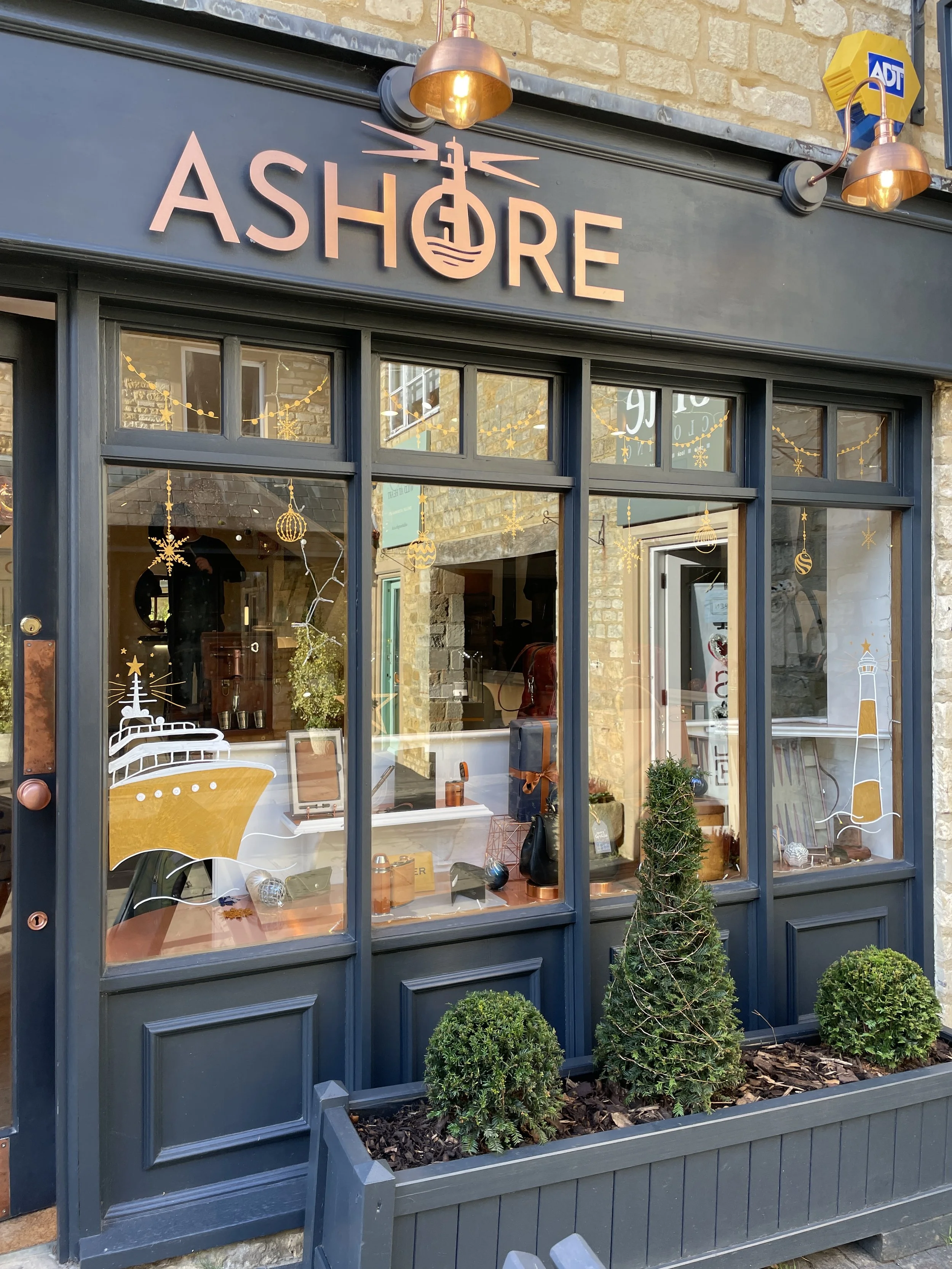 Exterior view of a storefront named 'ASHORE' with large windows decorated with Christmas ornaments, a potted plant, and Christmas-themed decals. There are two copper-colored outdoor lights and an ADT security sign on the wall.