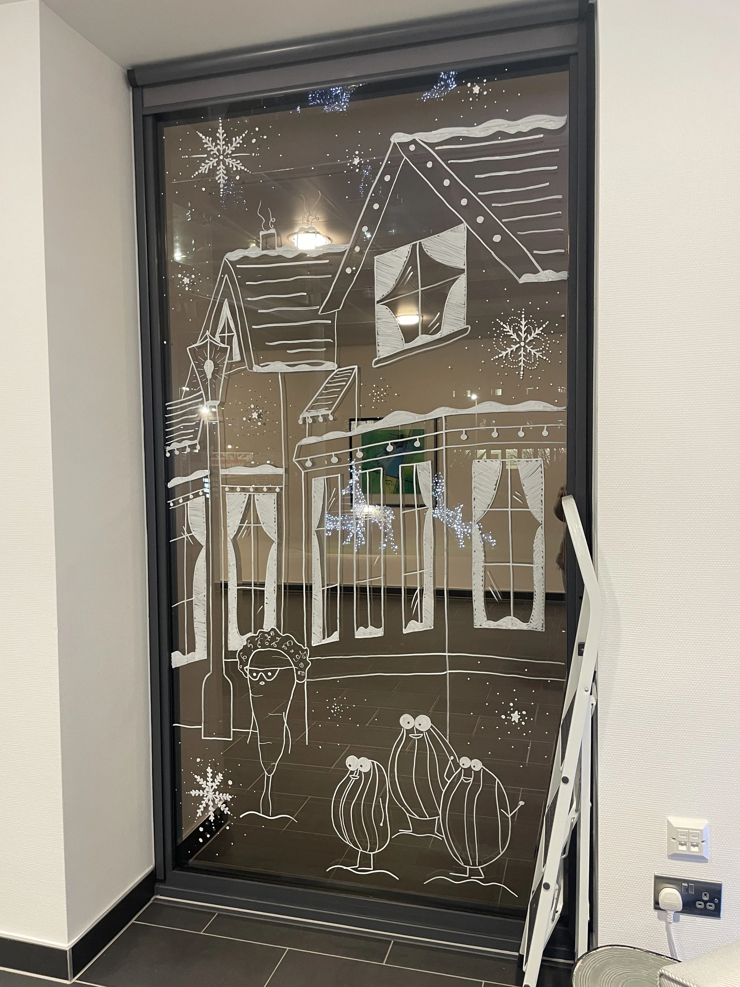 Decorative holiday window art featuring a house, snowflakes, a person with glasses, and three creatures with eyes, drawn in white on a glass window.