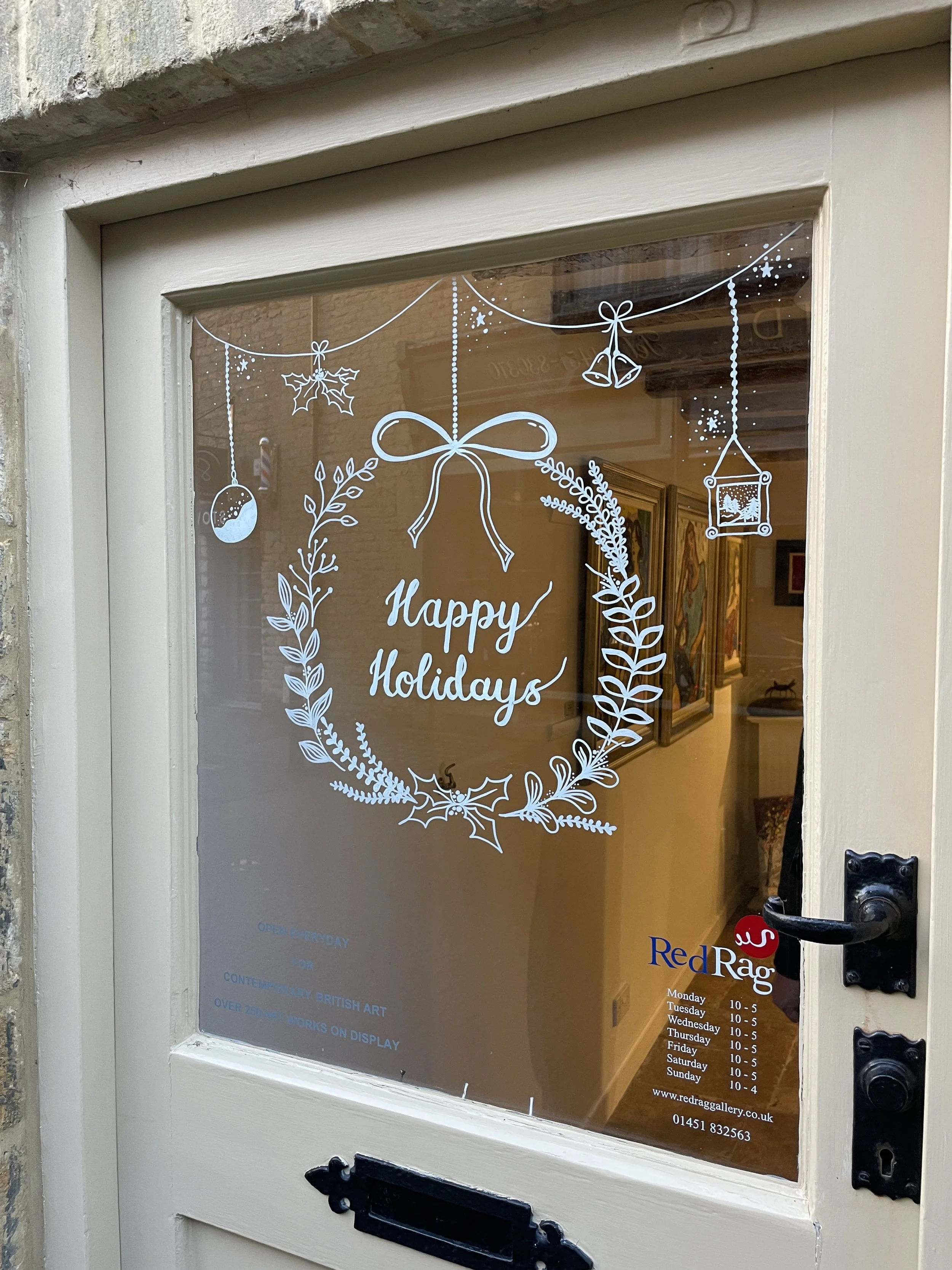 A glass door decorated with white holiday-themed illustrations including a wreath with the words "Happy Holidays," a bow, holly, and hanging ornaments. The inside of the building has framed artwork on the walls.