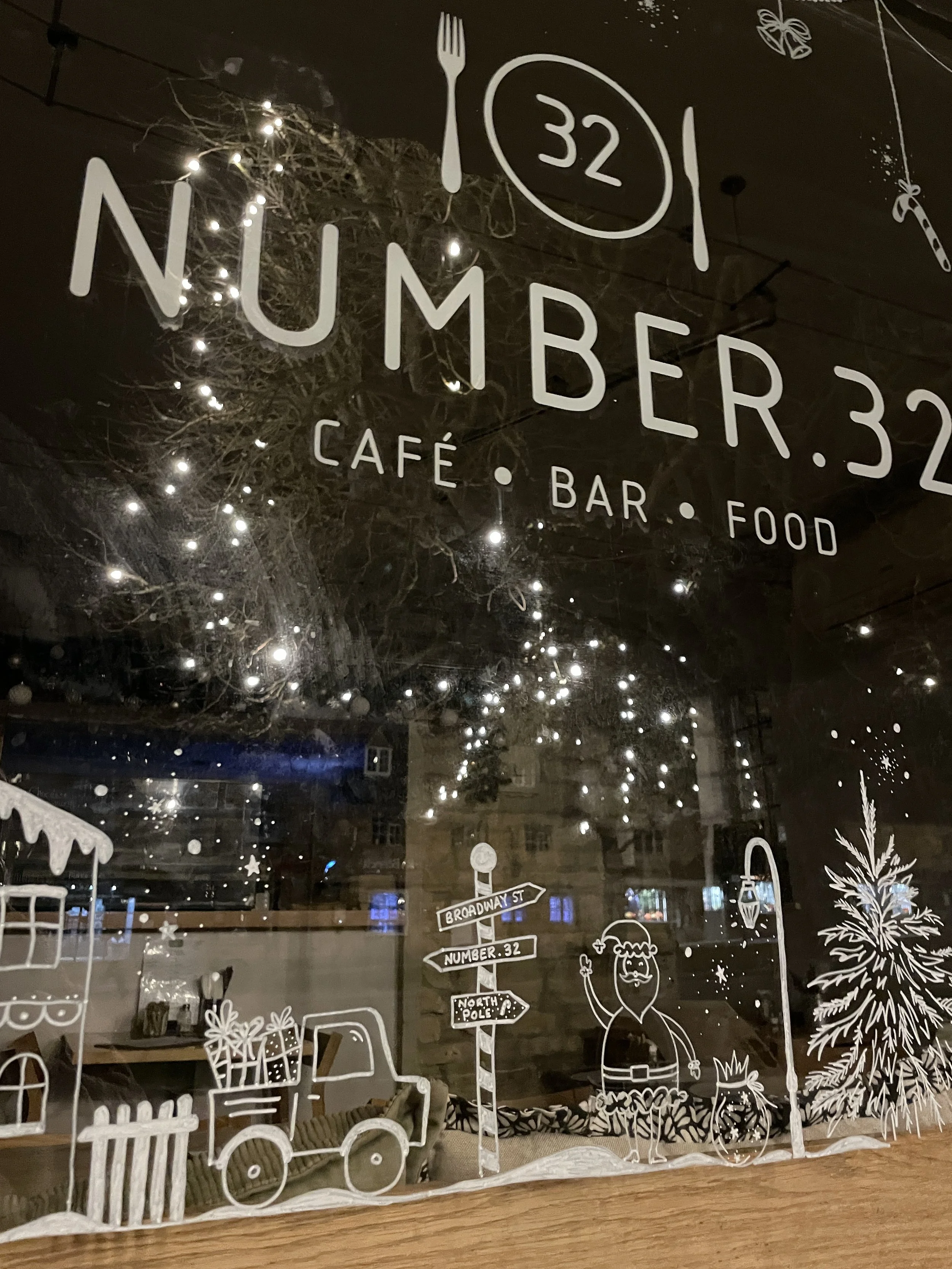 Decorative window with white illustrations of a Santa Claus, a Christmas tree, gift boxes, a street sign, and holiday lights, advertising café, bar, and food at Number 32 on Broadway Street.