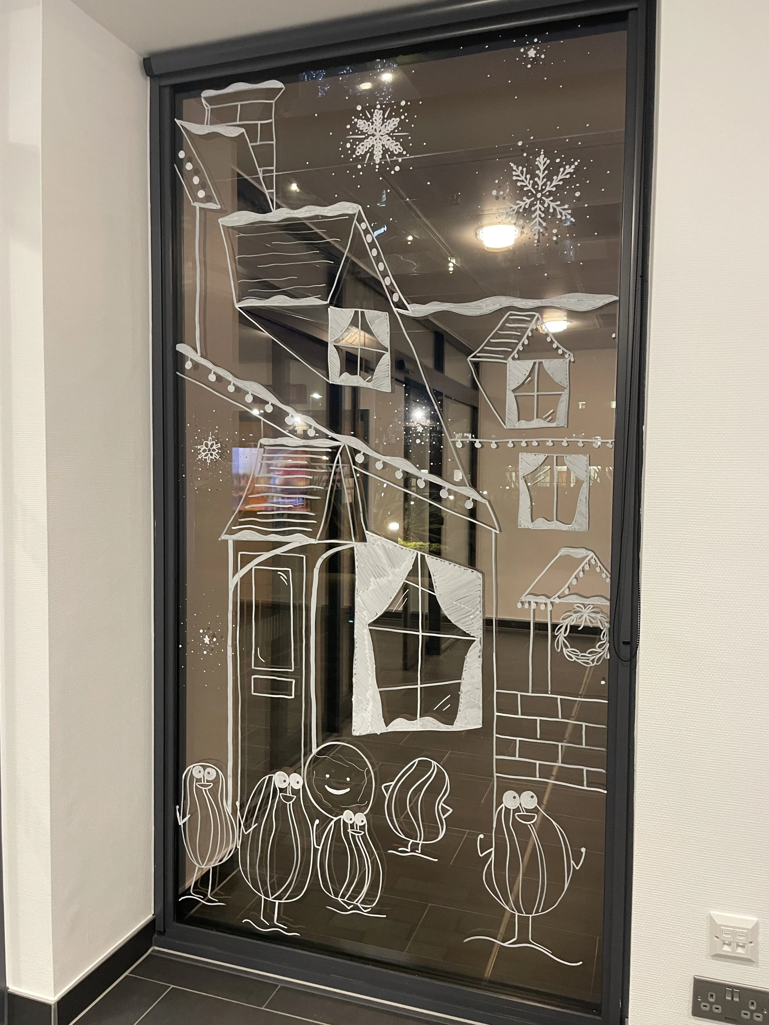 Window decorated with white illustrations of a house, hanging lanterns, snowflakes, and cartoon vegetables with smiling faces.