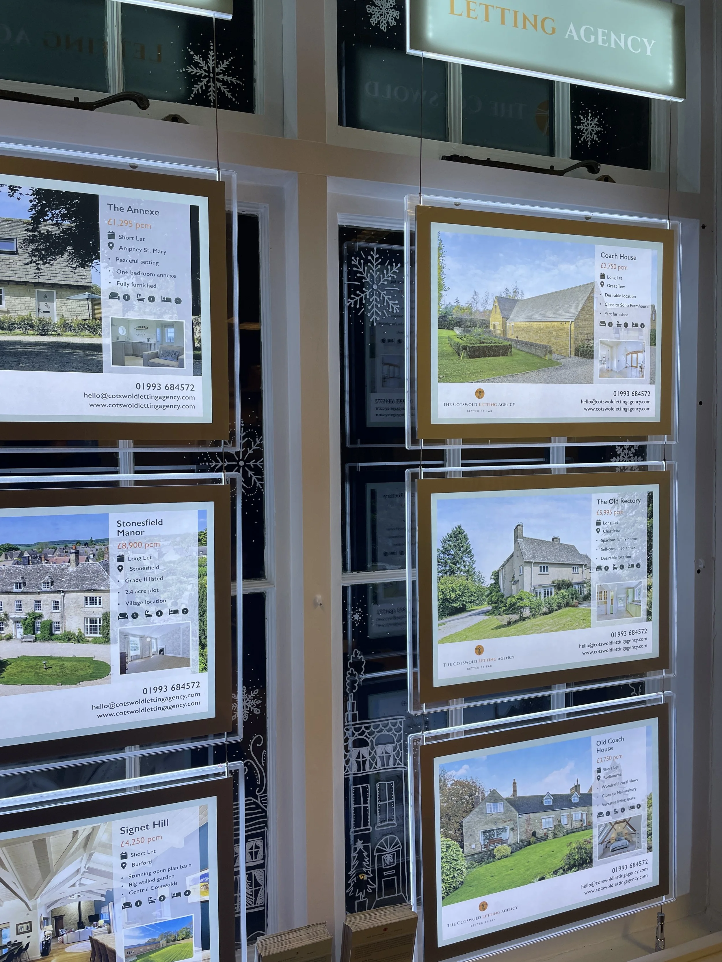 Display panels advertising various real estate properties for sale, including photos, prices, and descriptions, inside a window with decorative snowflakes.