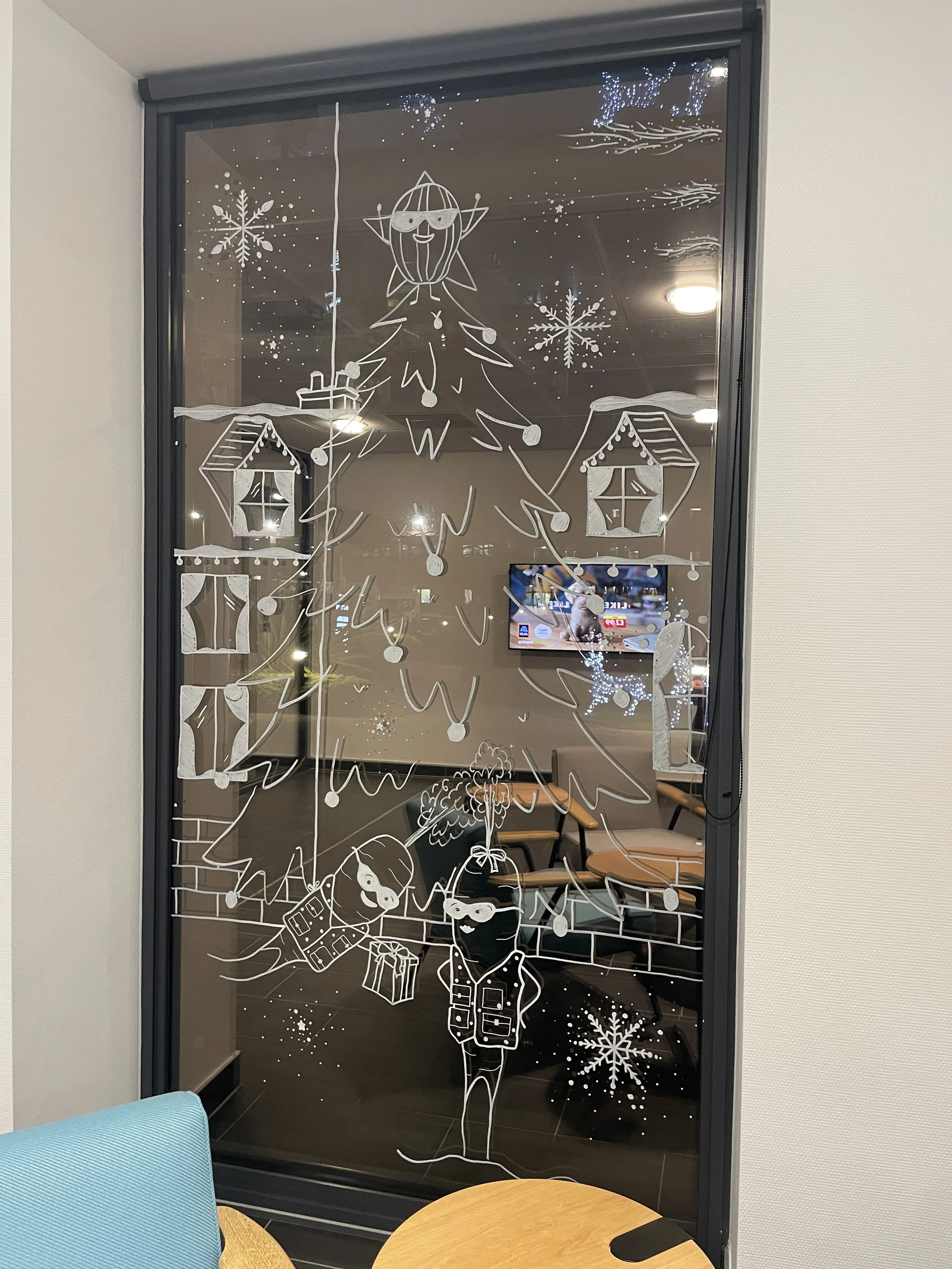 Decorative Christmas window art featuring a large Christmas tree with ornaments, a star on top, surrounded by snowflakes, houses, and two people in winter coats with presents, all sketched in white on the glass window.