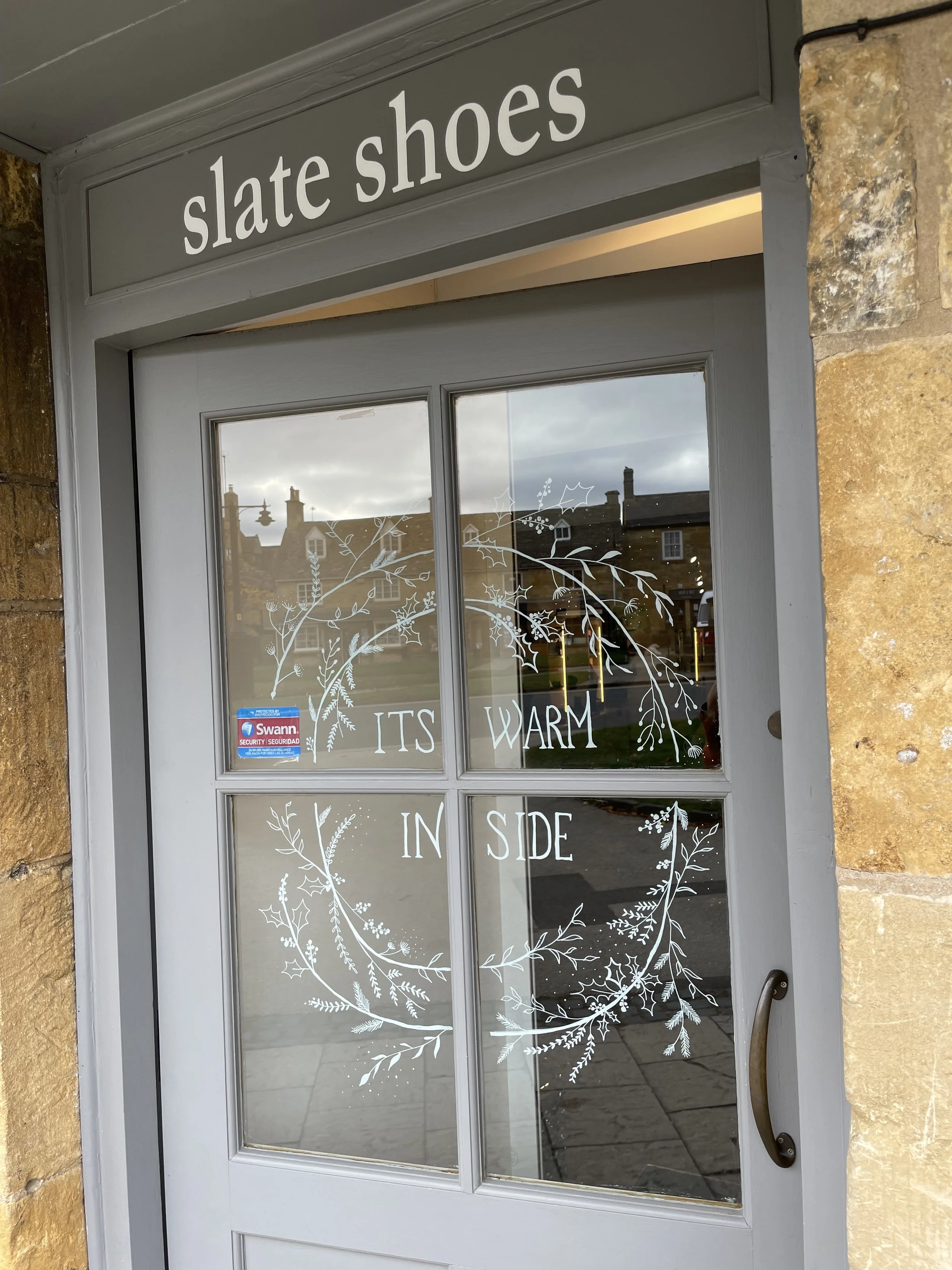 Storefront door with a sign that reads "slate shoes." The door has decorative white illustrations of branches and leaves, along with the words "IT'S WARM INSIDE."