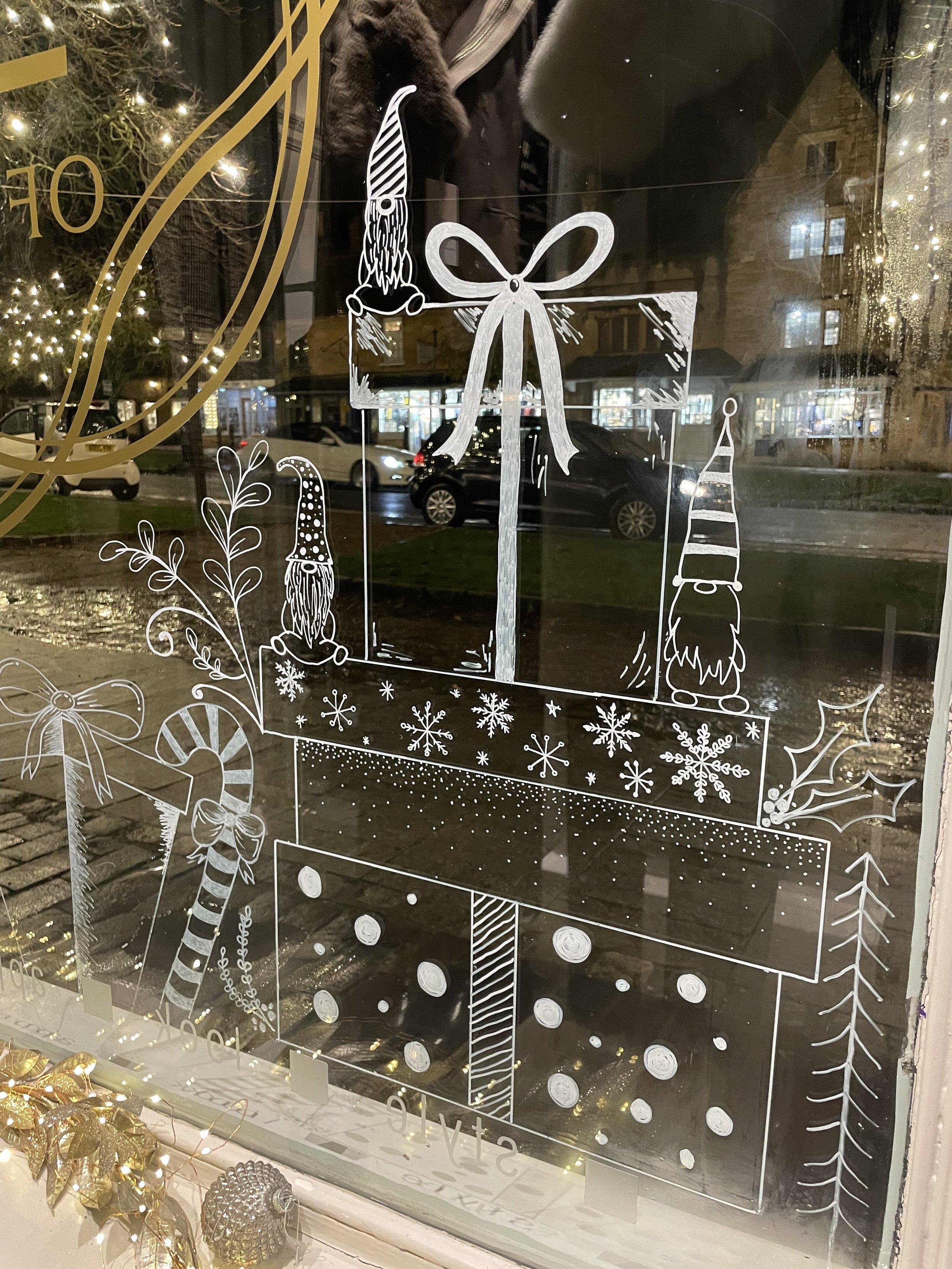 Decorative window with white line art of a Christmas gift box with a bow, surrounded by holiday elements such as holly, candy cane, snowflakes, Christmas gnomes, a Christmas tree, and fairy lights in the background.