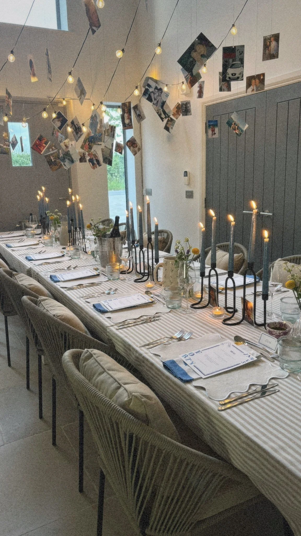 Long dining table set for a meal with candles, flowers, and place settings, decorated with hanging photos and string lights.