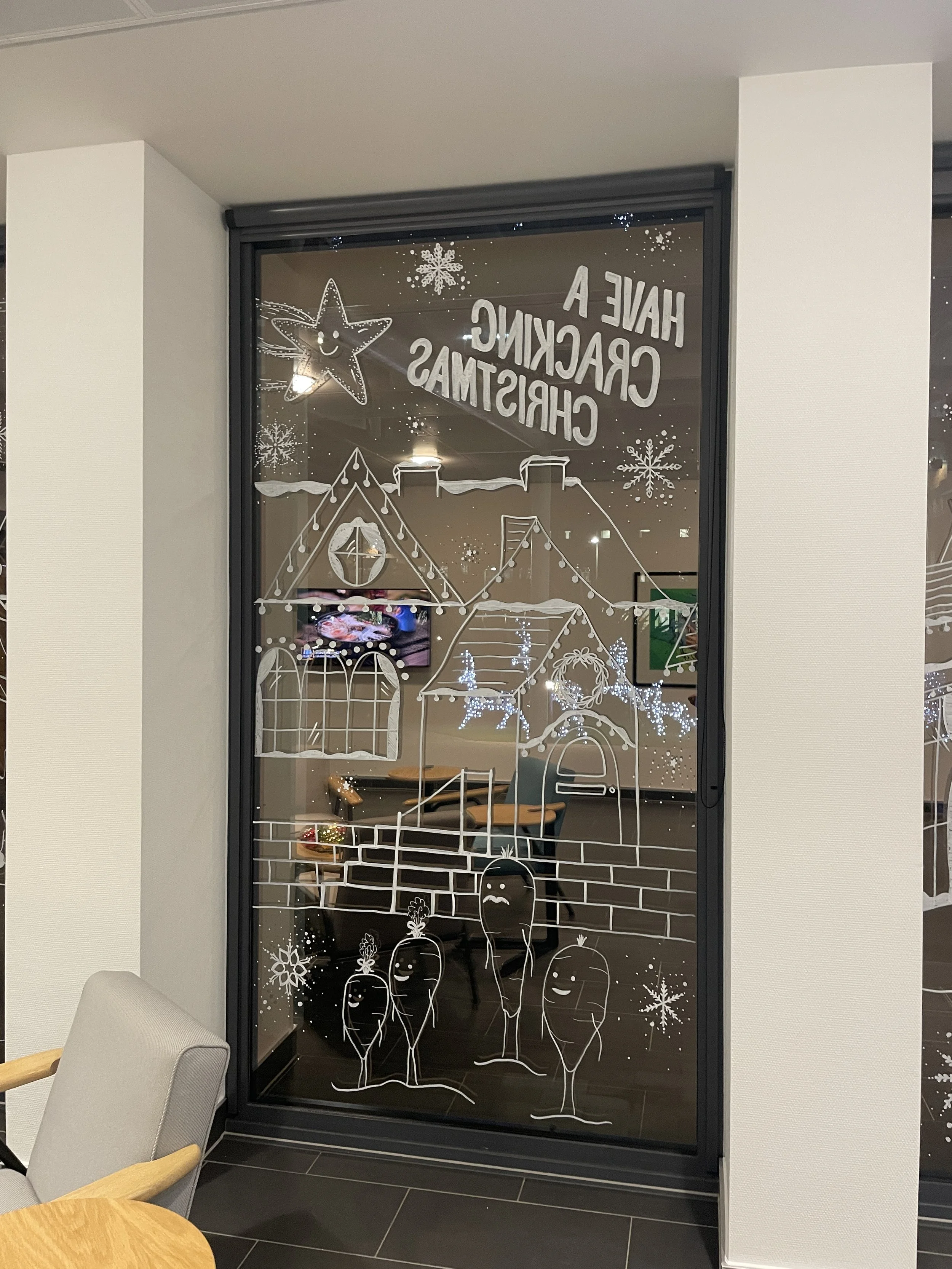 Window with holiday-themed white illustrations of a house, snowflakes, a smiling star, and four carrots with happy faces, along with the message 'Have a Cracking Christmas'.