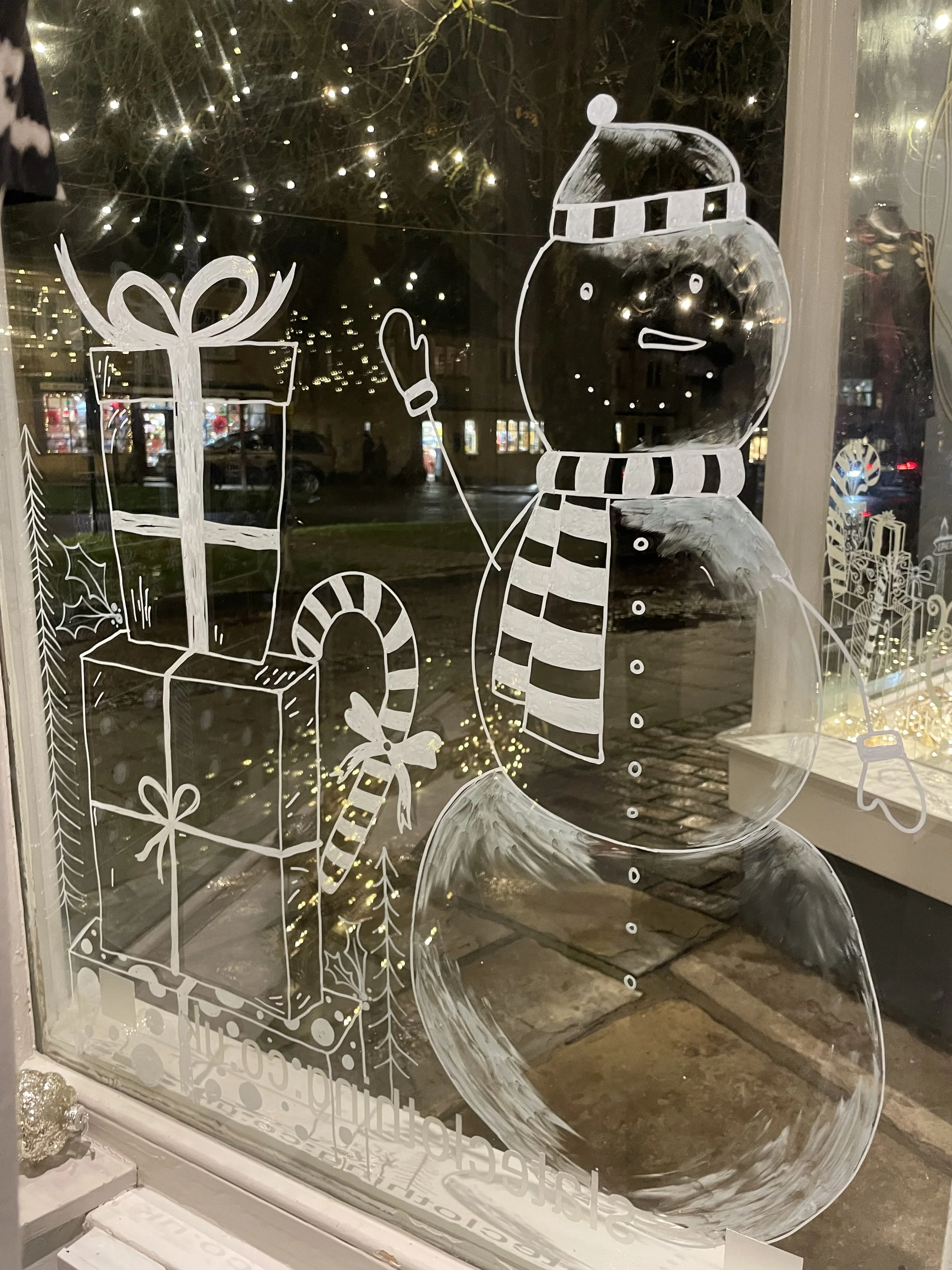 Window with a hand-painted snowman wearing a striped scarf and hat, holding a mitten in a winter scene with Christmas presents and holiday decorations reflected outside.