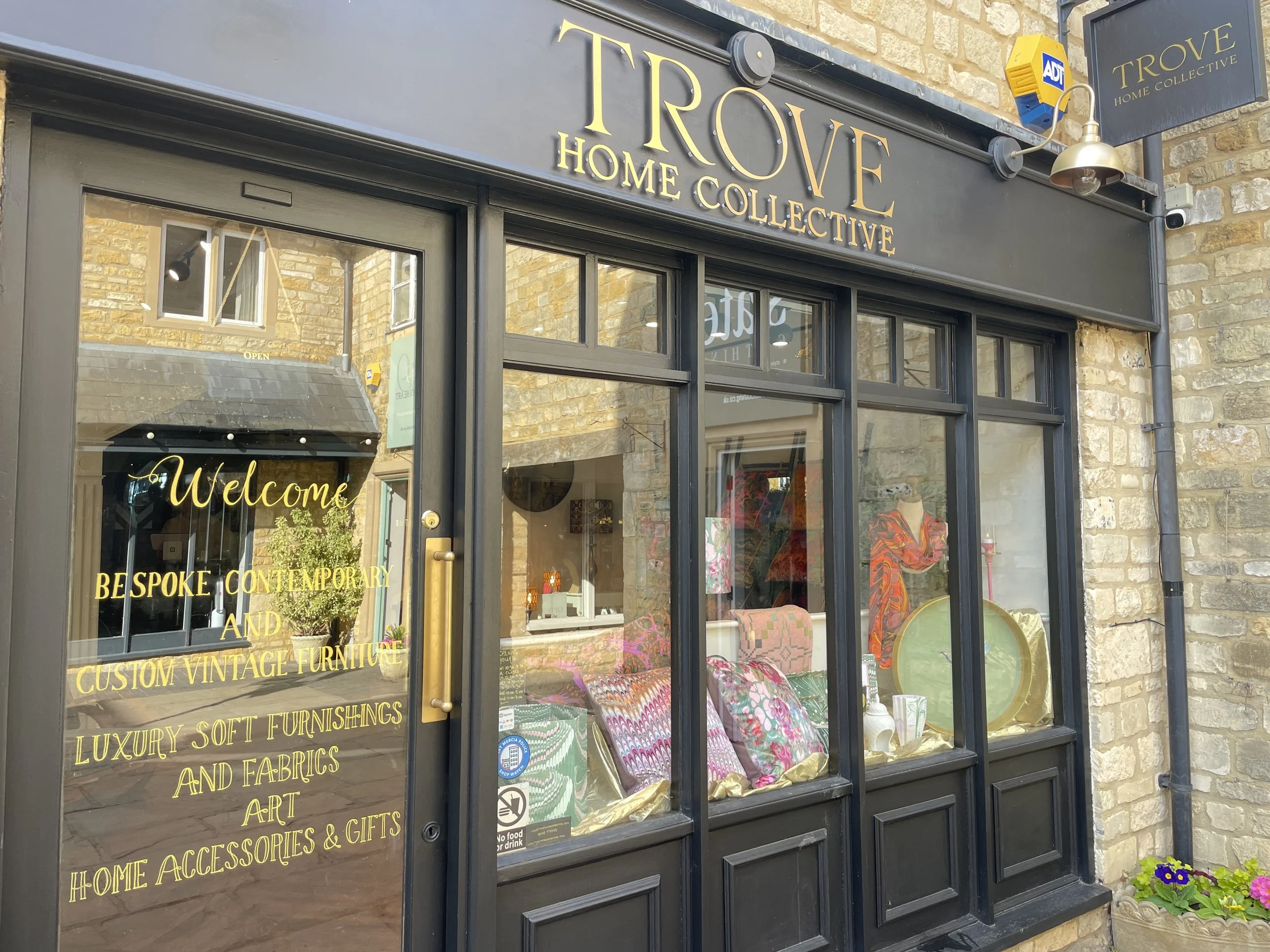 Exterior of a store called 'Trove Home Collective' with large glass windows displaying colorful furniture and decor, including cushions, trays, and art pieces. The shop has a black facade with gold lettering, and the glass door has a welcome message 