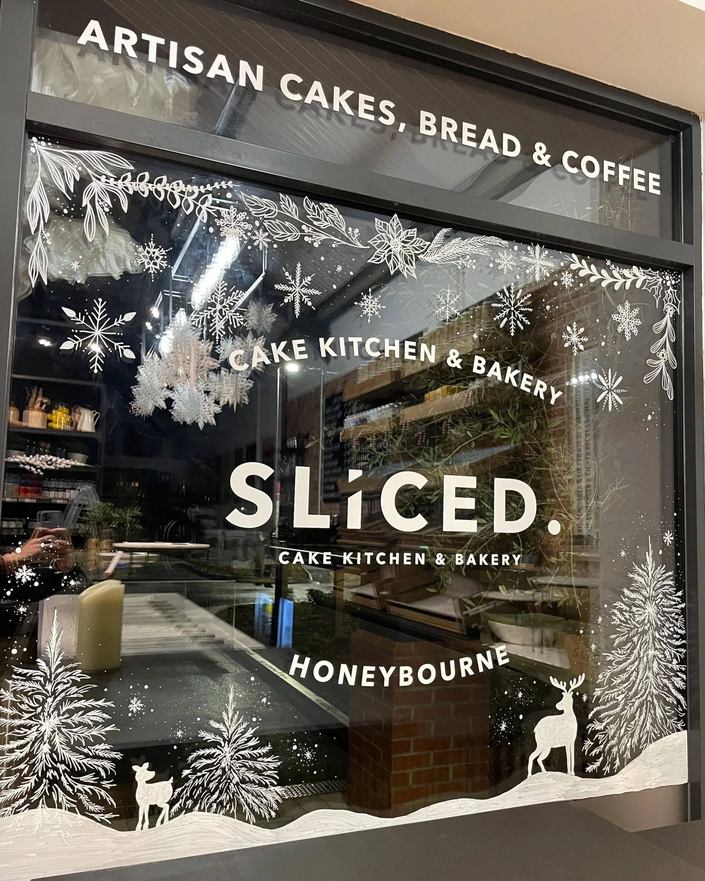 A glass window with white winter-themed decorations including snowflakes, pine trees, and a reindeer, advertising a bakery called 'Cake Kitchen & Bakery' that offers artisan cakes, bread, and coffee, with the word 'SLICED' prominently displayed.