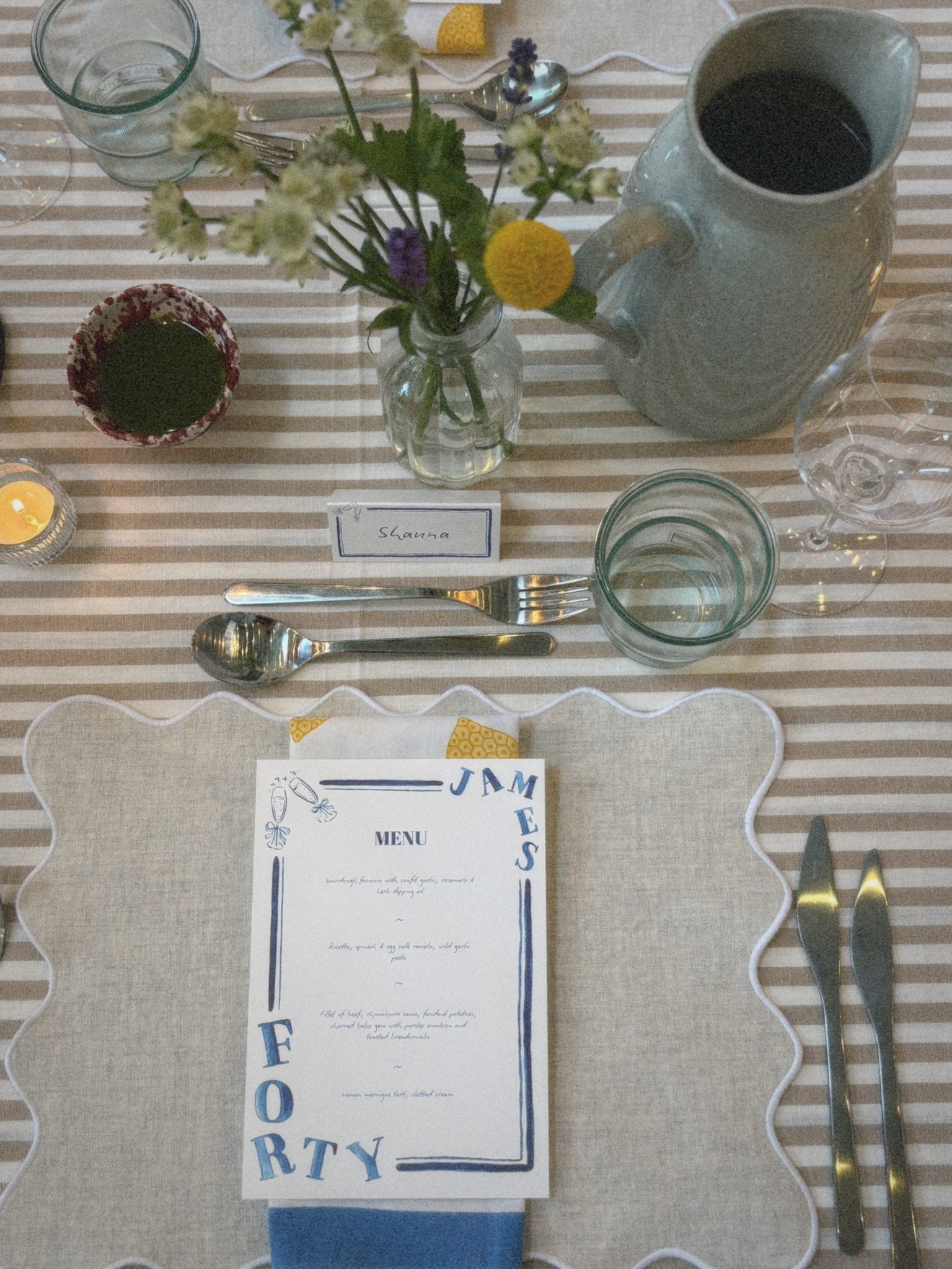 Table setting with a menu, flowers in a vase, a glass of water, a large tea pot, a small bowl of dark sauce, cutlery, a small candle, and a place card labeled 'Shanna', on a striped tablecloth.