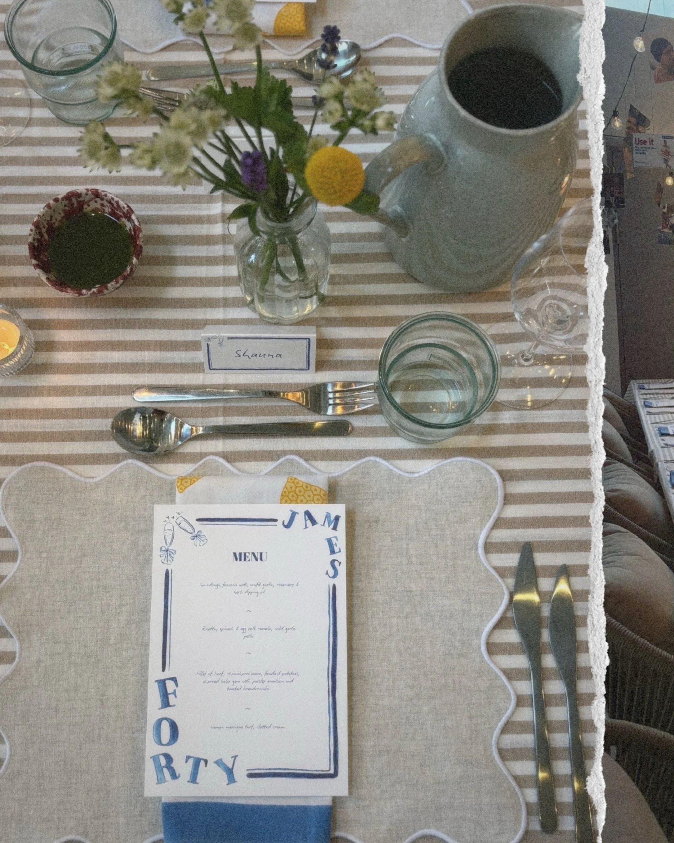 A table setting with a striped tablecloth, a small vase of flowers, a water pitcher, glasses, a small bowl, utensils, a menu card, and a place card with the name 'Shanna'.