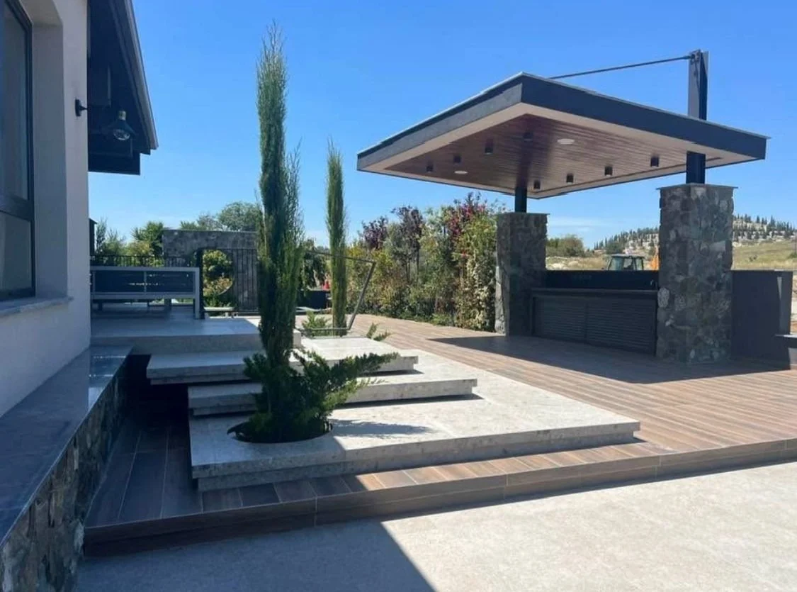Outdoor patio area with a stone and wooden structure, tall cypress trees, and landscaped plants under a clear blue sky.