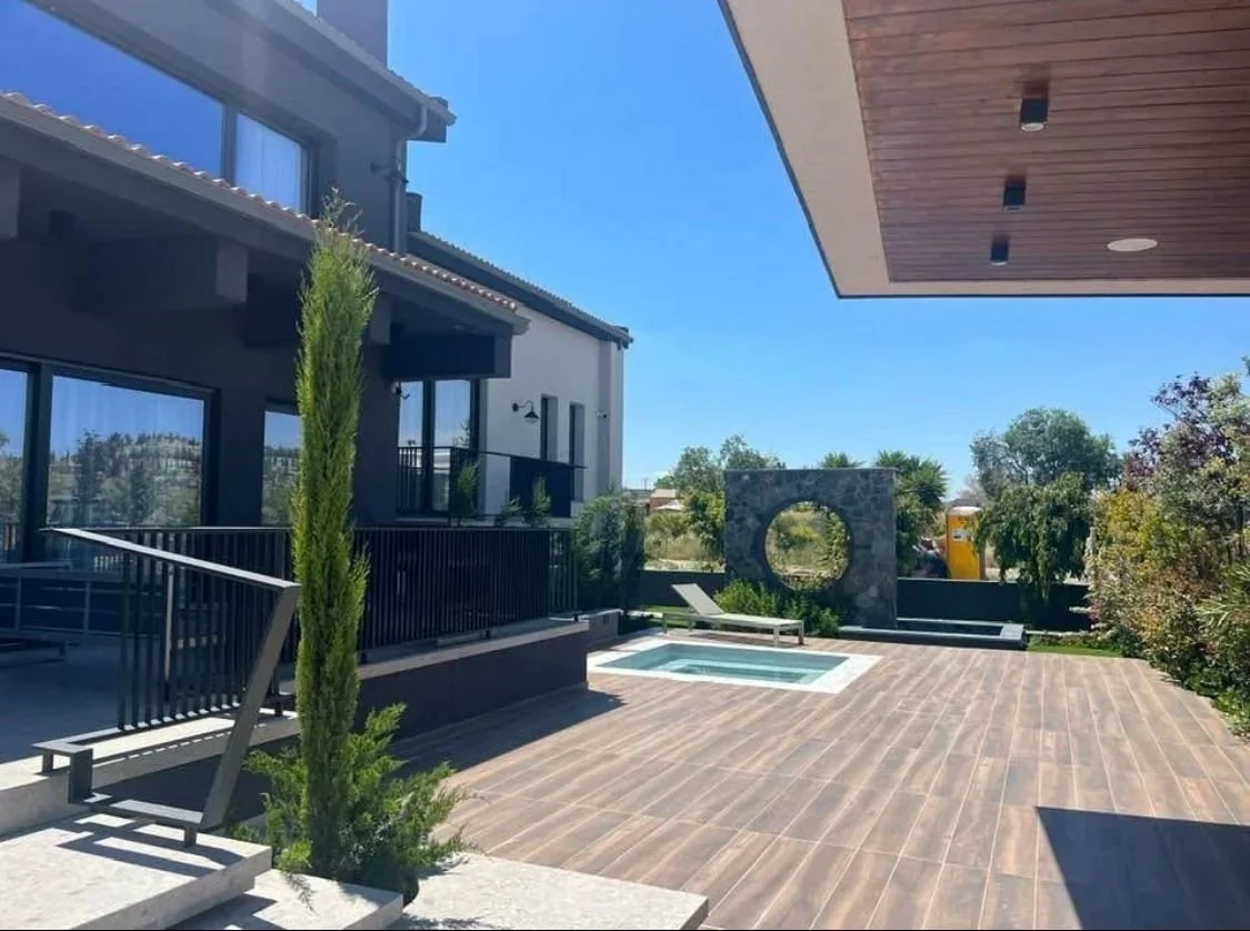 Modern outdoor patio area with a small pool, wooden deck, and a stone wall with a circular cutout, surrounded by greenery and a clear blue sky.
