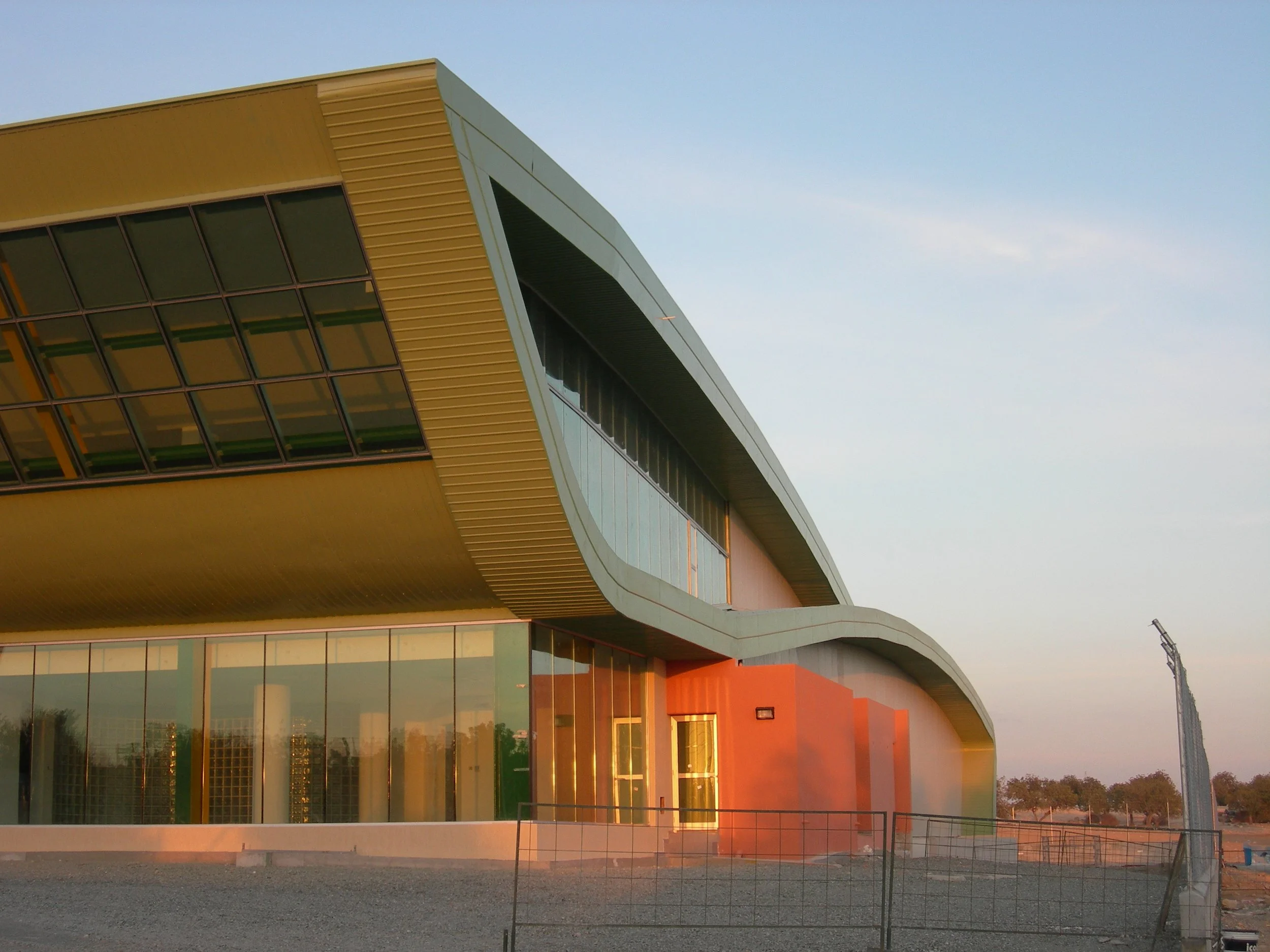 Modern building with curved design and glass windows, illuminated by the setting sun, with a fence and open landscape in the foreground.