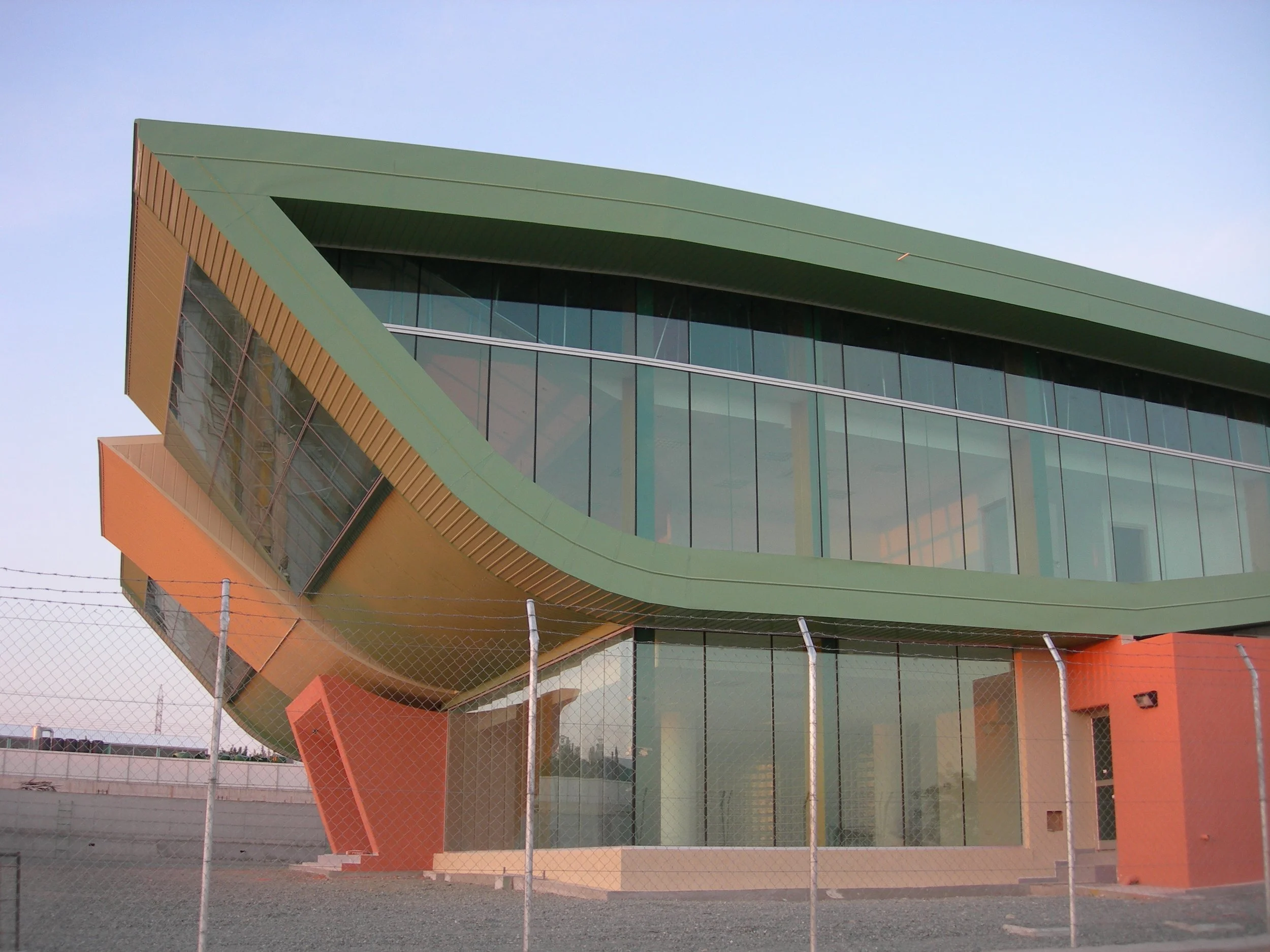 Modern building with angular design, large glass windows, green and orange exterior panels, and a chain-link fence in the foreground.