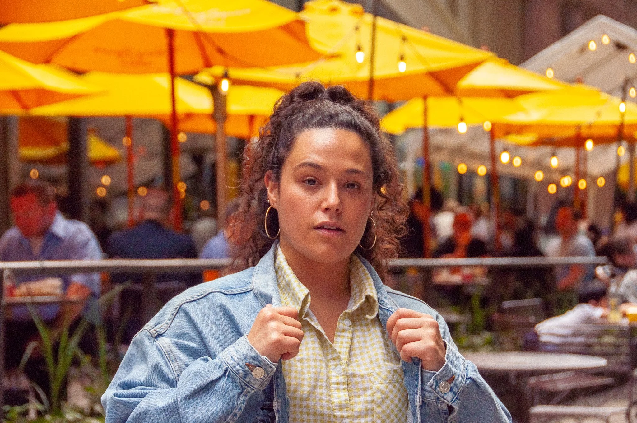 A woman with curly hair, wearing hoop earrings, a yellow checkered shirt, and a denim jacket, standing outdoors at a cafe with yellow umbrellas and string lights in the background.