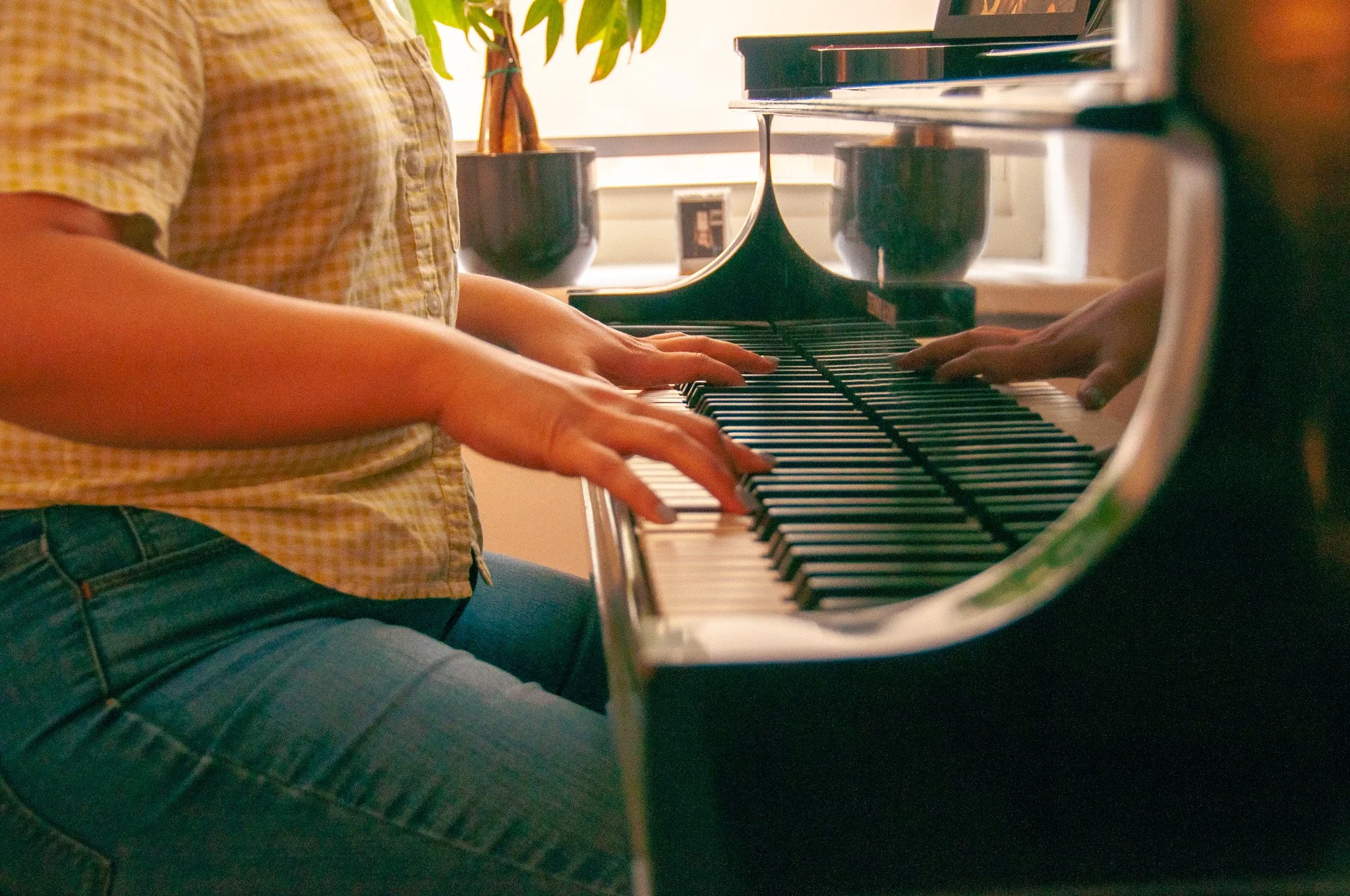 Two individuals playing a black grand piano together, one wearing a yellow checkered shirt, in a room with potted plants and sunlight coming through a window.