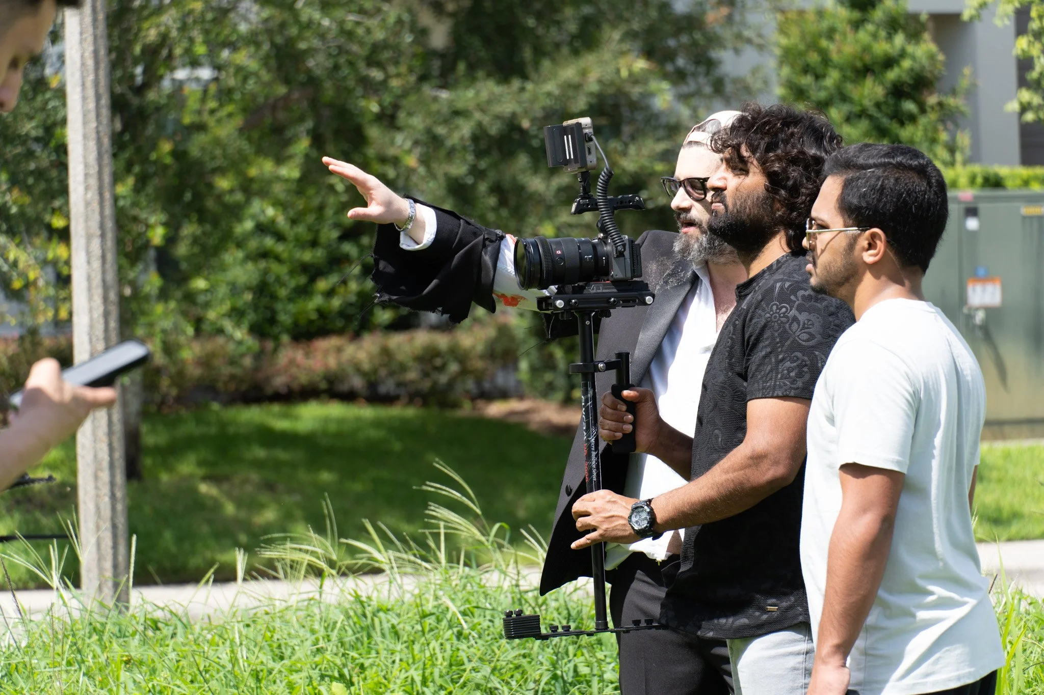 Two men standing outdoors, looking at a camera one of them is holding, against a backdrop of a yellow wall, wooden fence, and a small green shrub.