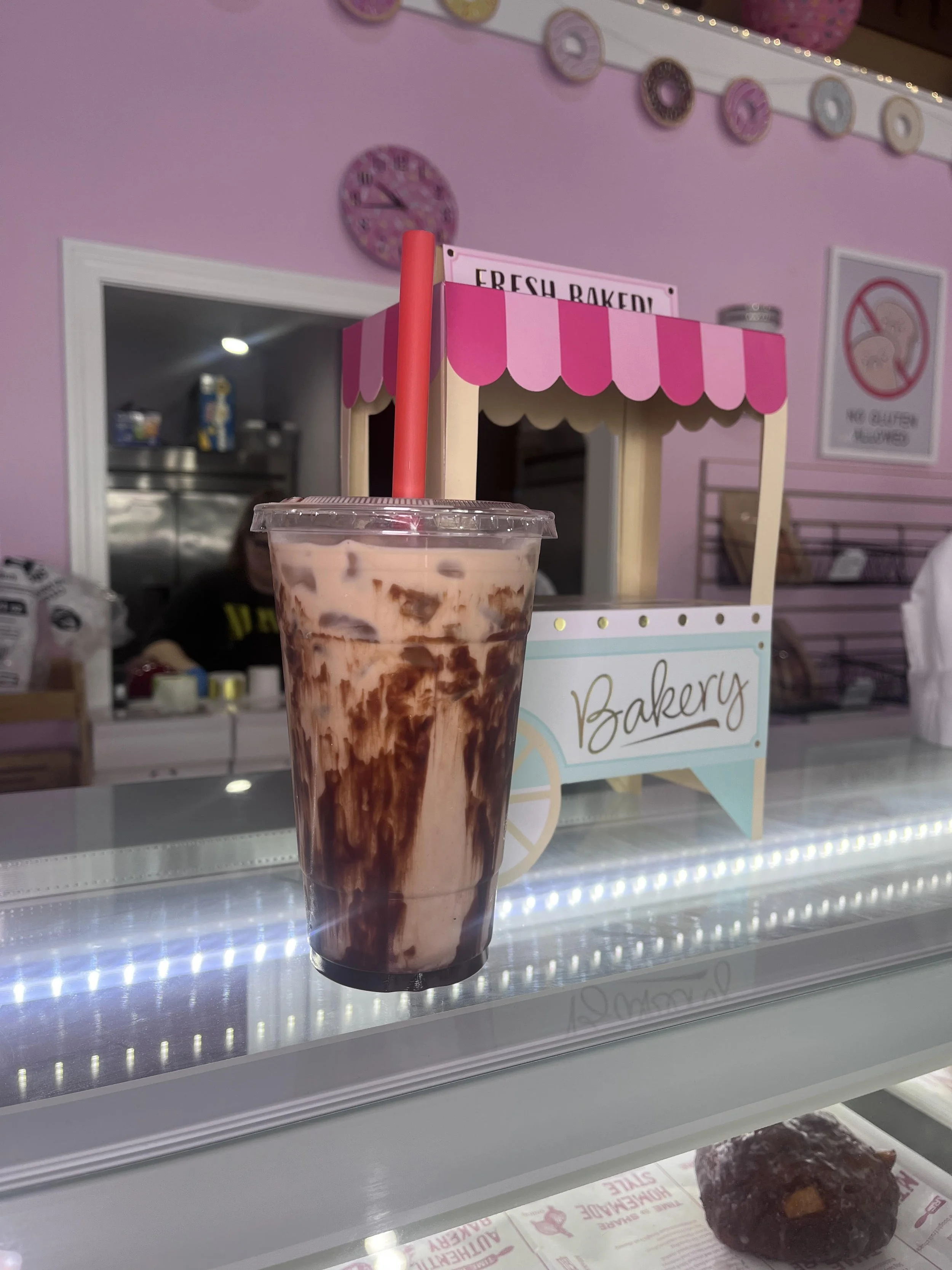 Chocolate coffee with chocolate syrup in a clear cup with a pink straw, standing on a glass display case in a bakery with pink walls and bakery signage.