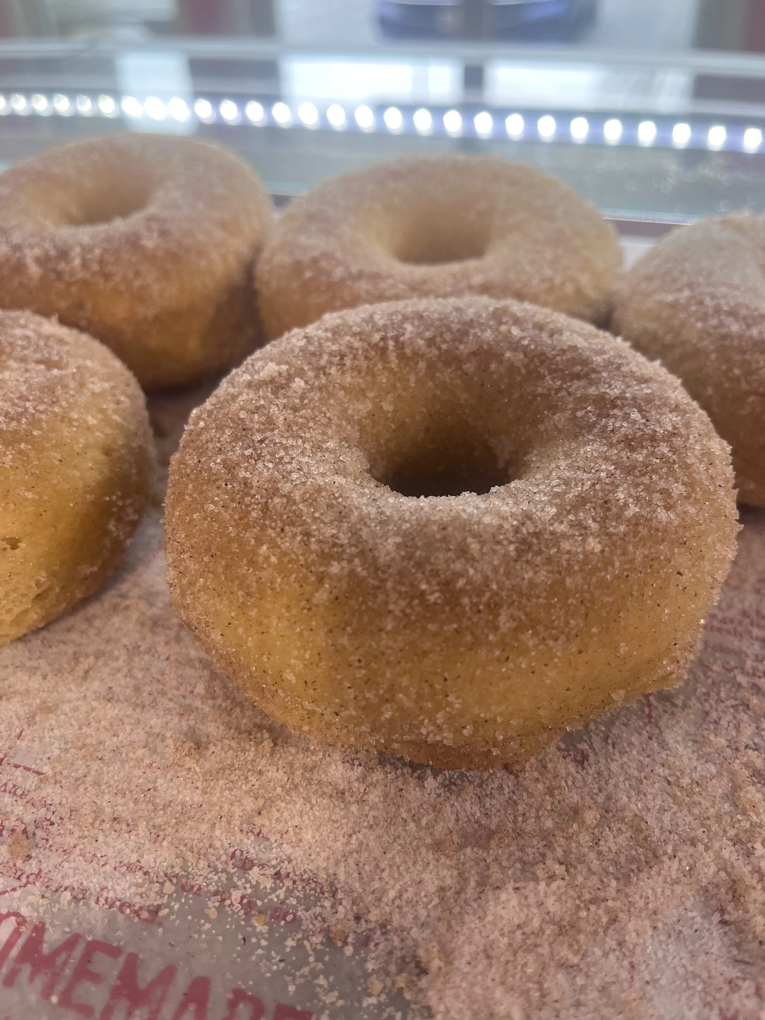 Close-up of sugar-coated donuts on a paper wrapper with some pink text, in a display case. Gluten free