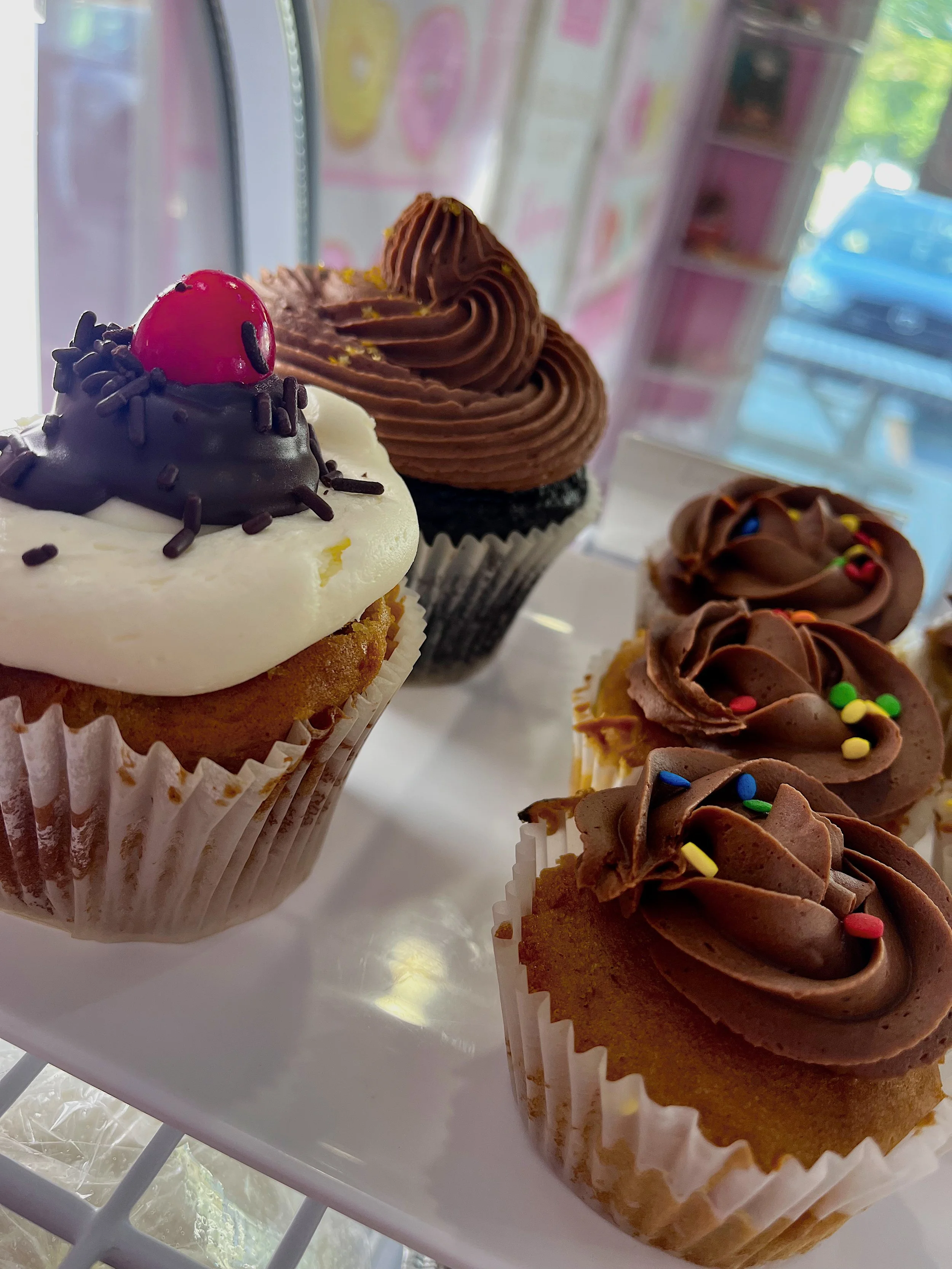 Assorted cupcakes with chocolate and vanilla frosting, sprinkles, and decorative toppings on a white display stand inside a bakery.