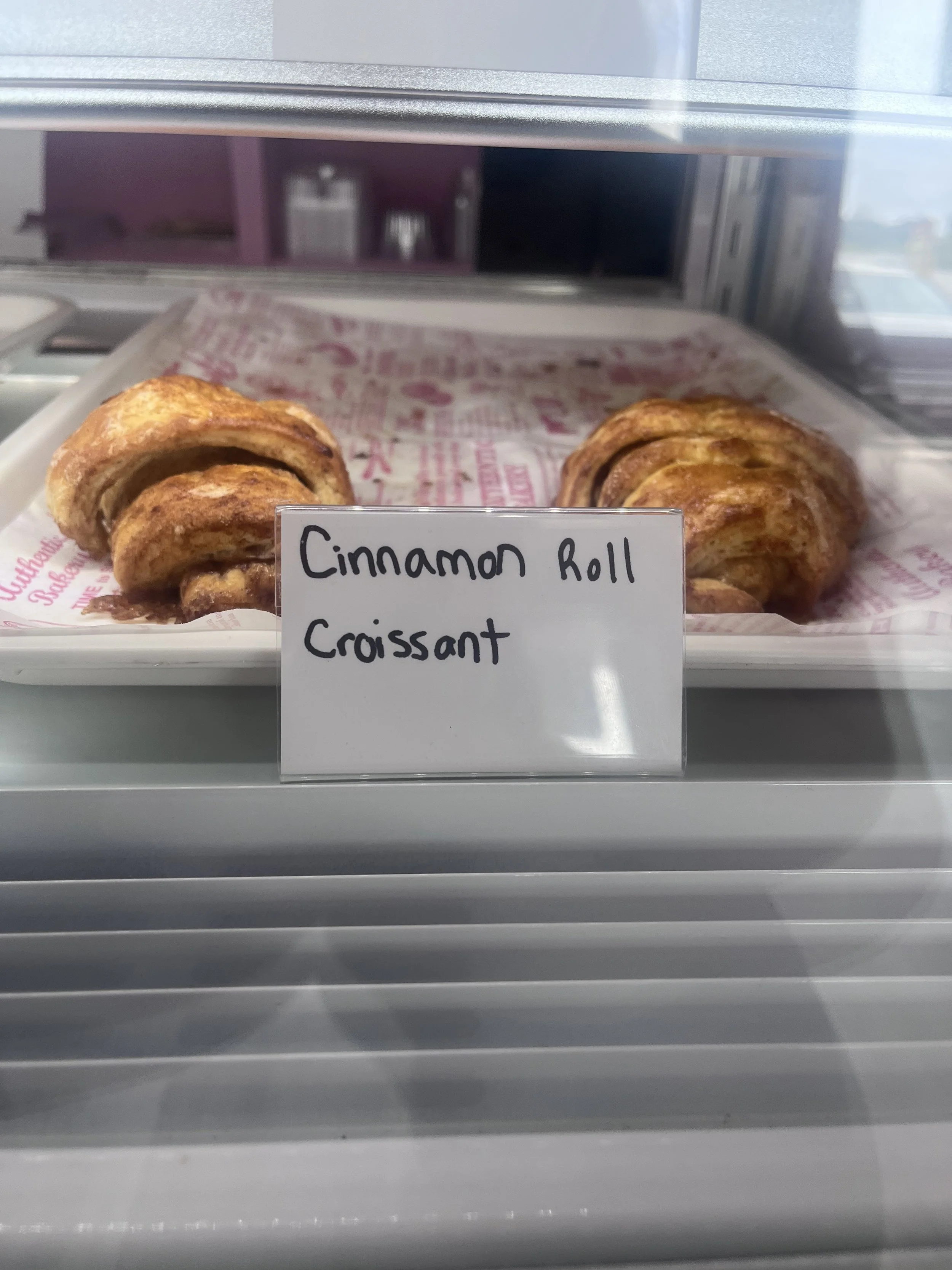 Cinnamon roll croissant displayed on a tray in a bakery case with a sign that reads 'Cinnamon Roll Croissant'.