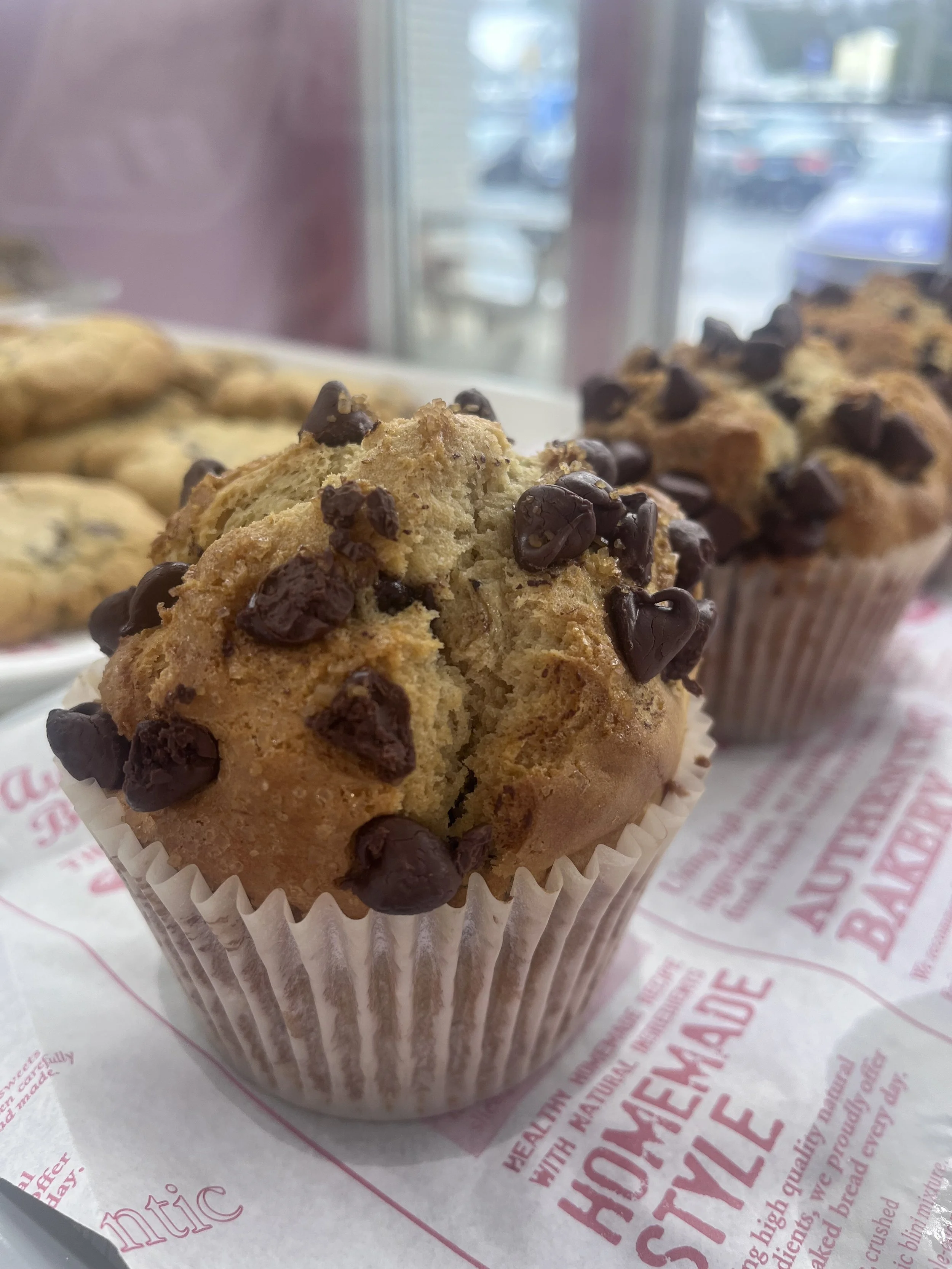 Chocolate chip muffin in a paper wrapper with a second muffin in the background on paper with red text, near a window revealing a parking lot outside.