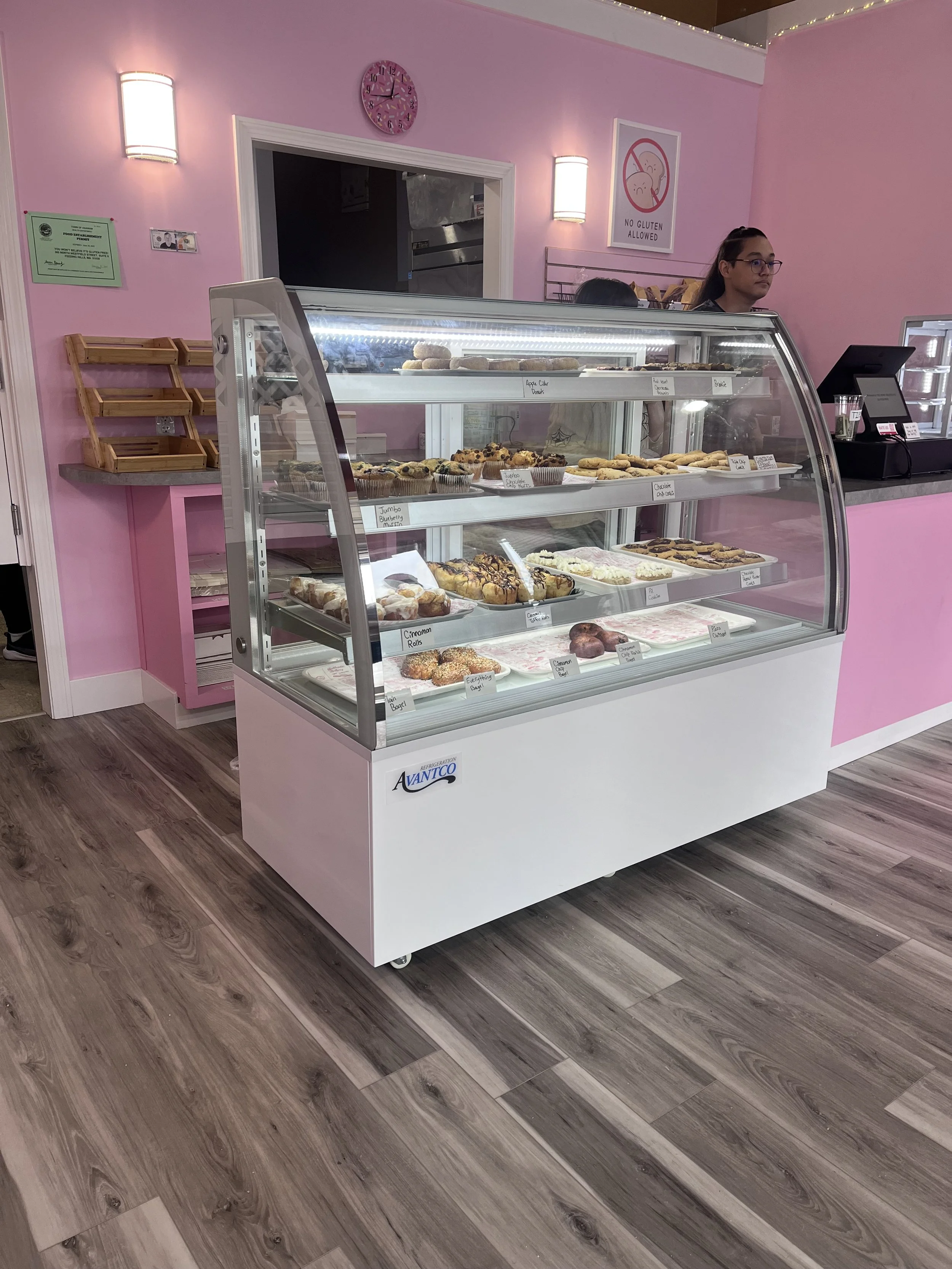 Display case with assorted baked goods such as cookies, muffins, and pastries inside a pink bakery with pink walls, a clock, and signs on the wall.