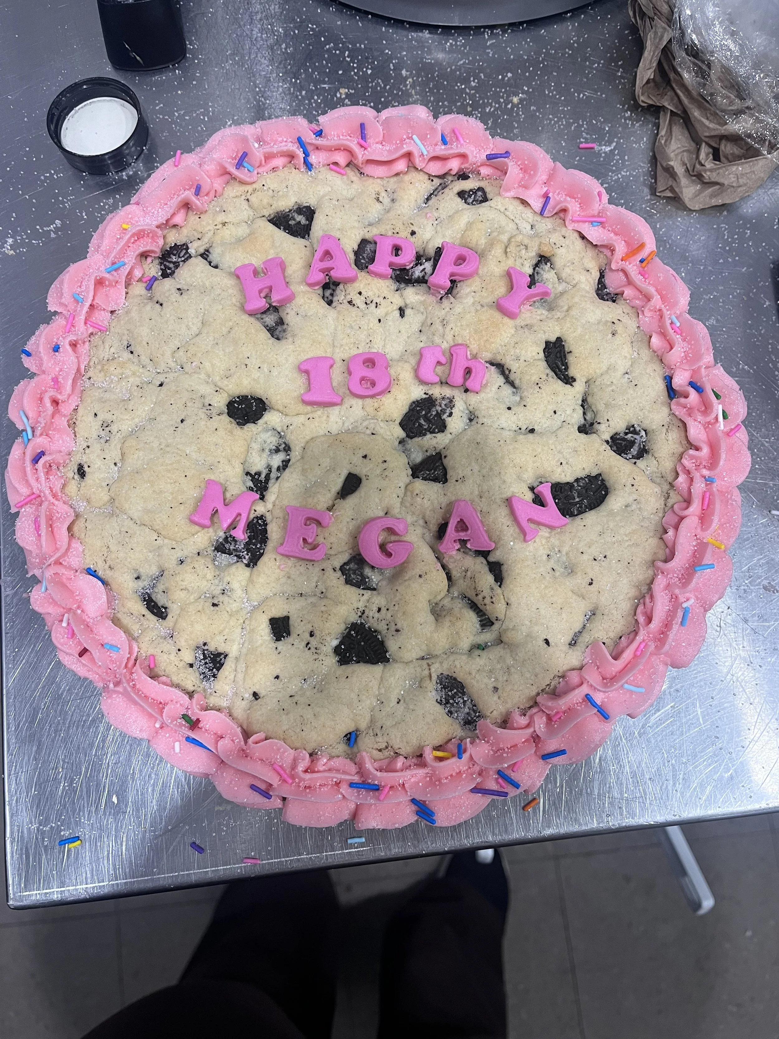 A large cookie cake decorated with pink frosting and colorful sprinkles, with pink letters spelling out "HAPPY 18TH MEGAN" on top.