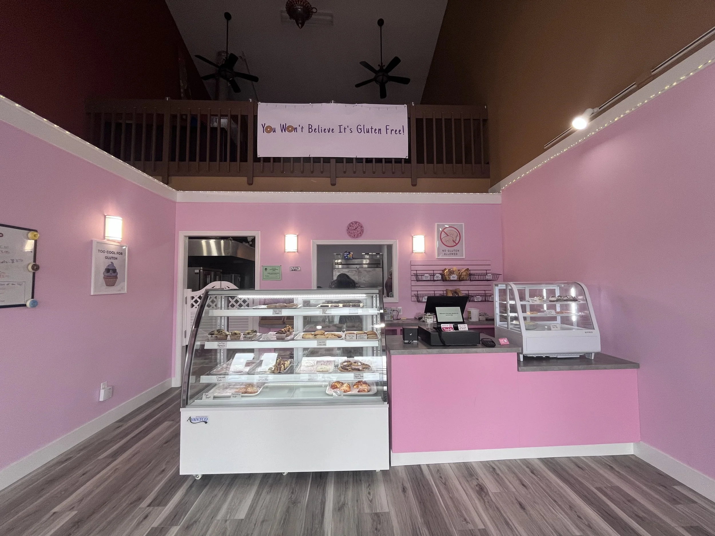 Interior of a bakery or cafe with pink walls, a glass display case containing pastries, a cash register, and a sign that reads "You Won't Believe It's Gluten Free!" on the upper level balcony.
