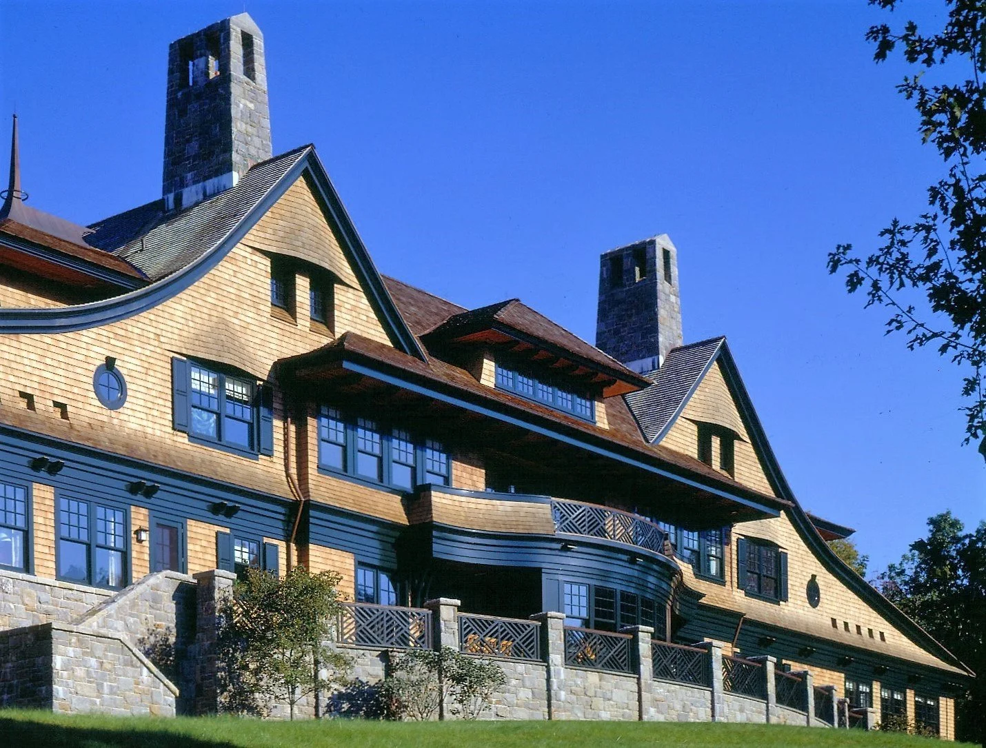 A large house with a wooden exterior, multiple gabled roofs, a stone foundation, and a spacious balcony, set against a bright blue sky.
