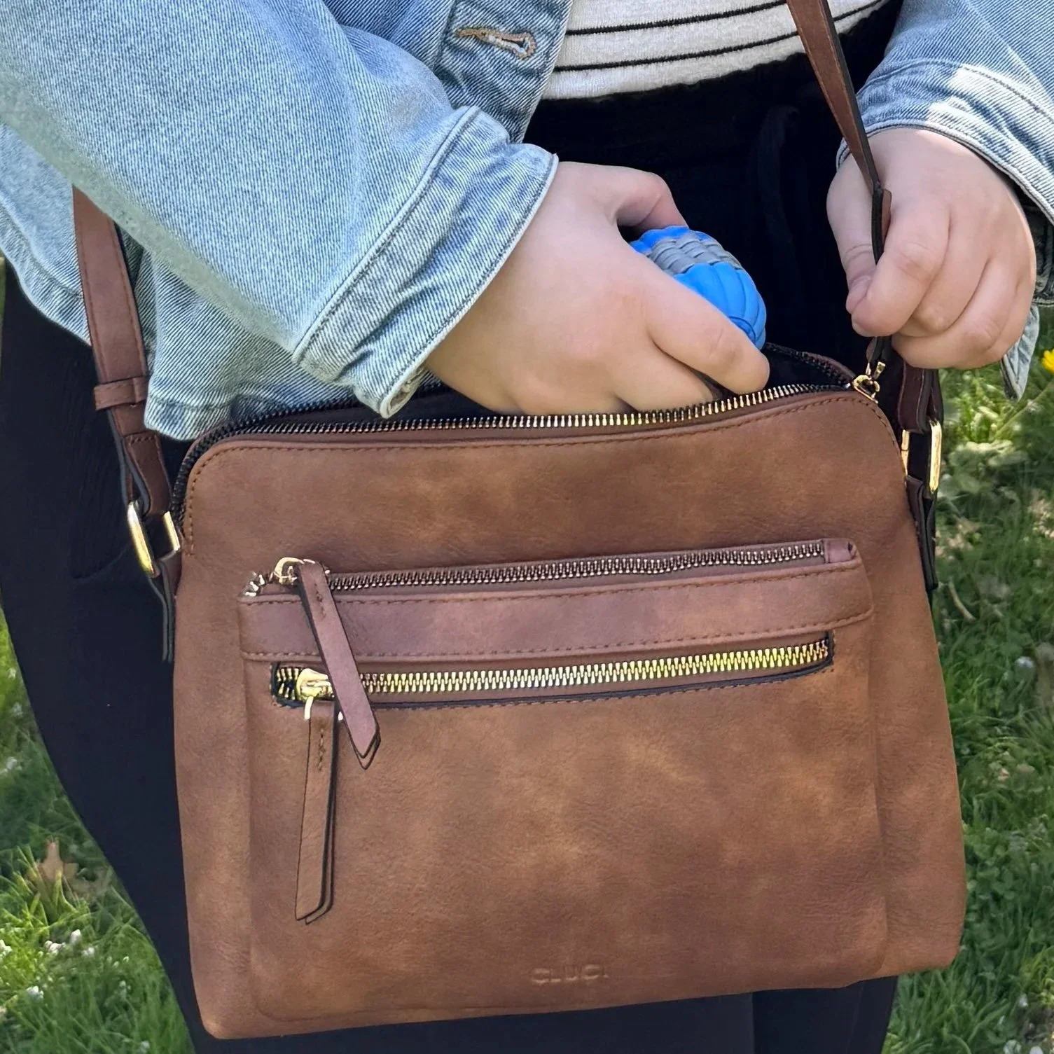 Person reaching into a brown leather bag with a blue and gray bubble-shaped object inside, outdoors with grass and small plants in the background.