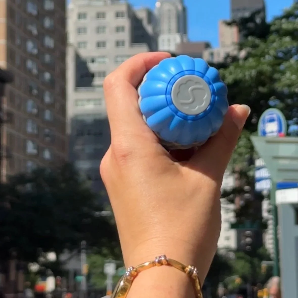 Hand holding a blue bubble toy against an urban cityscape background with tall buildings and trees.