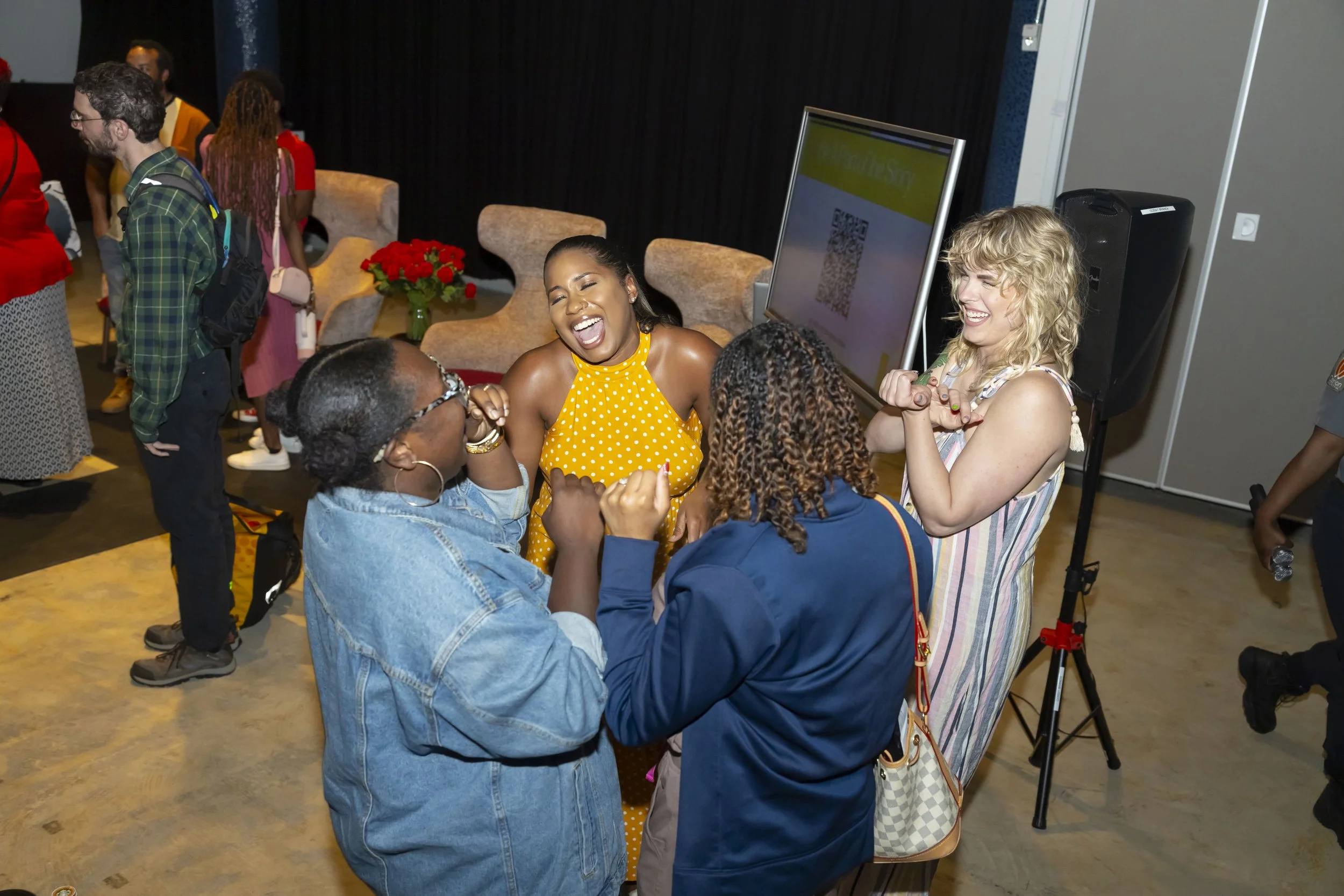 Group of women dancing and laughing together at an indoor event with chairs, a large screen, and people in the background.