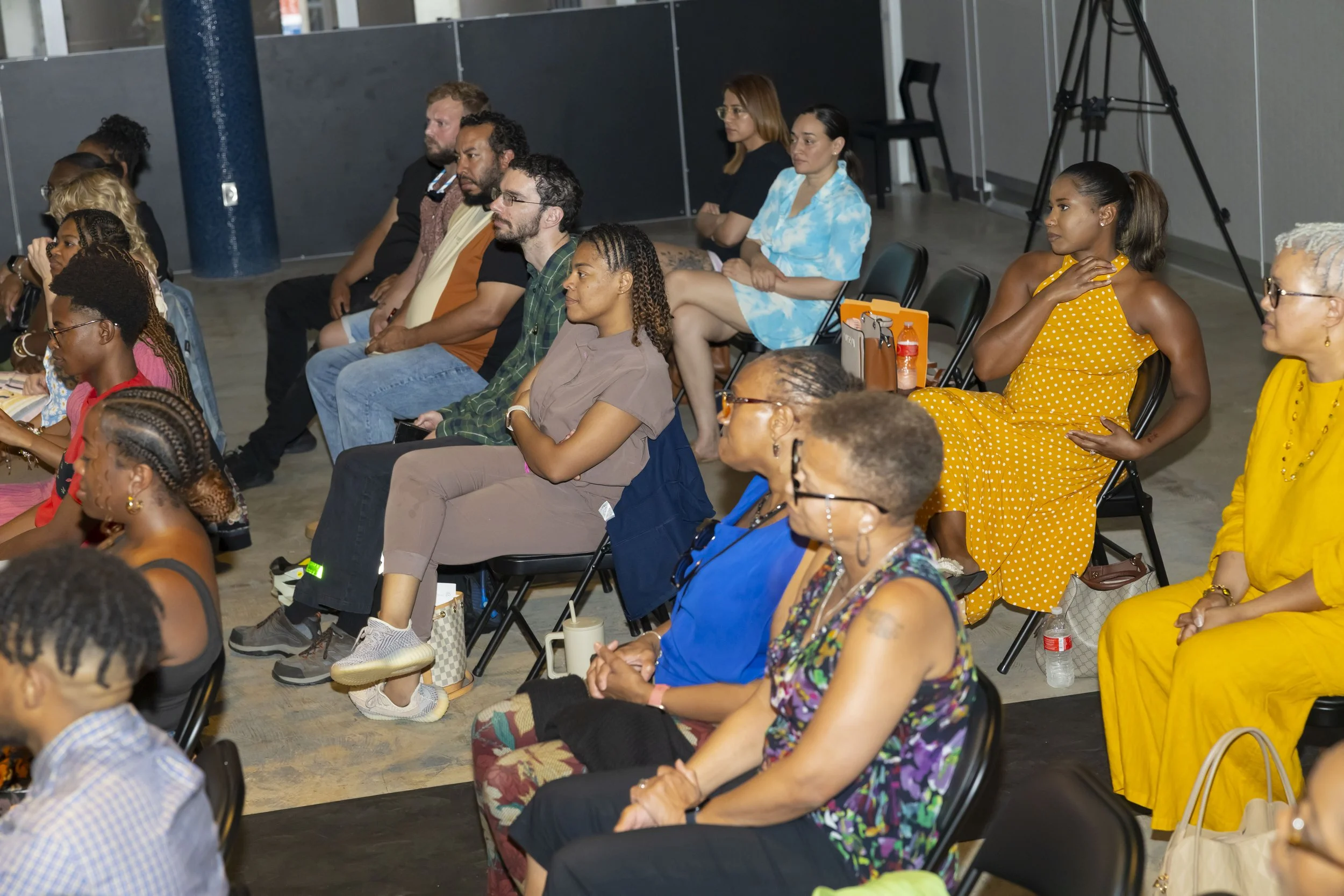 A group of people sitting on black chairs in an indoor event space, attentively listening to a presentation or speaker. The group includes diverse individuals, some wearing colorful clothing, and a camera setup is visible in the background.