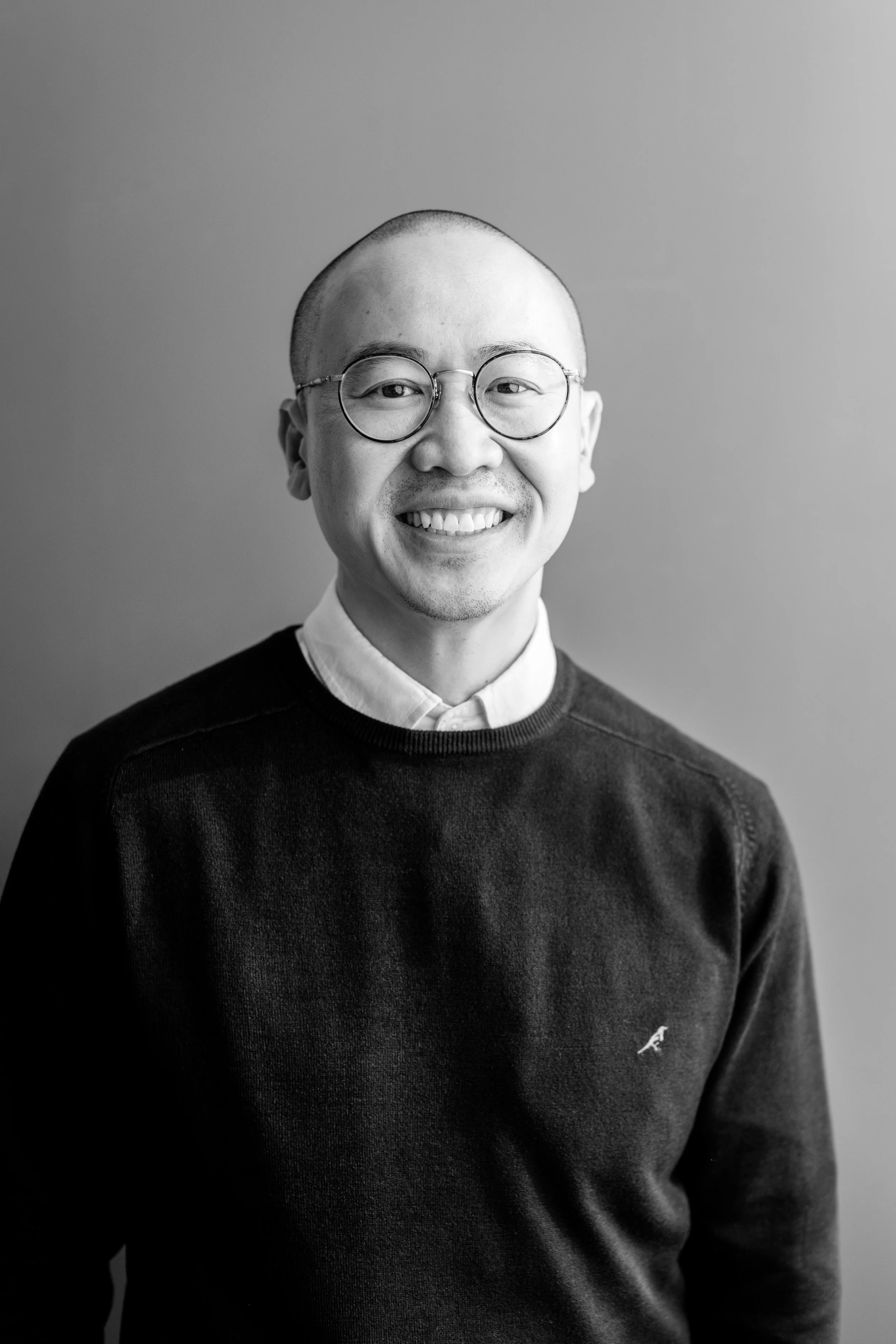 A smiling man with a shaved head and round glasses wearing a collared shirt against a plain background.