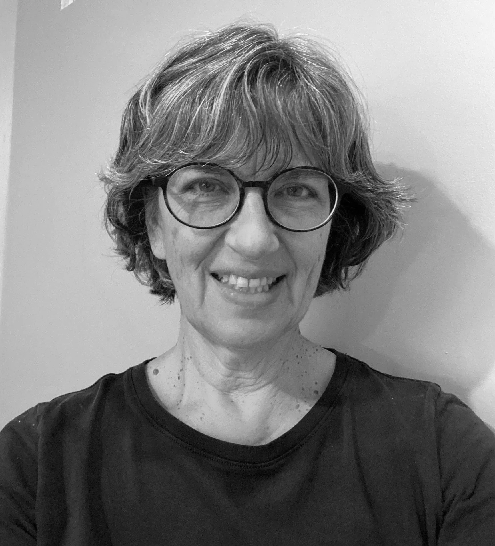 A woman with short, wavy hair wearing glasses and a black shirt, smiling in front of a plain wall.