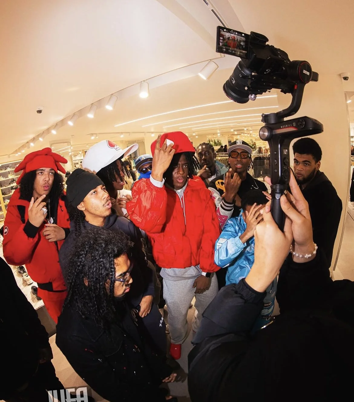 Viver and a group of people gathered around a camera in a shopping mall, some wearing colorful jackets and hats, with one person in the center adjusting a red jacket and cap.