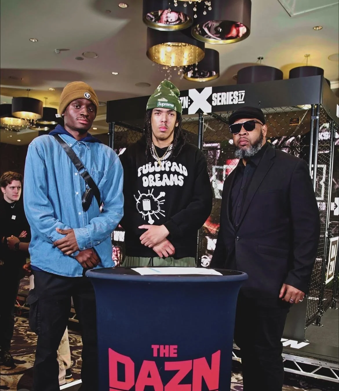 Vicer and two men standing behind a black table with a pink and blue banner that says "THE DAZN" at an event. Vicer in the middle is wearing a black hoodie with white writing and graphics, a green beanie, and jewelry.