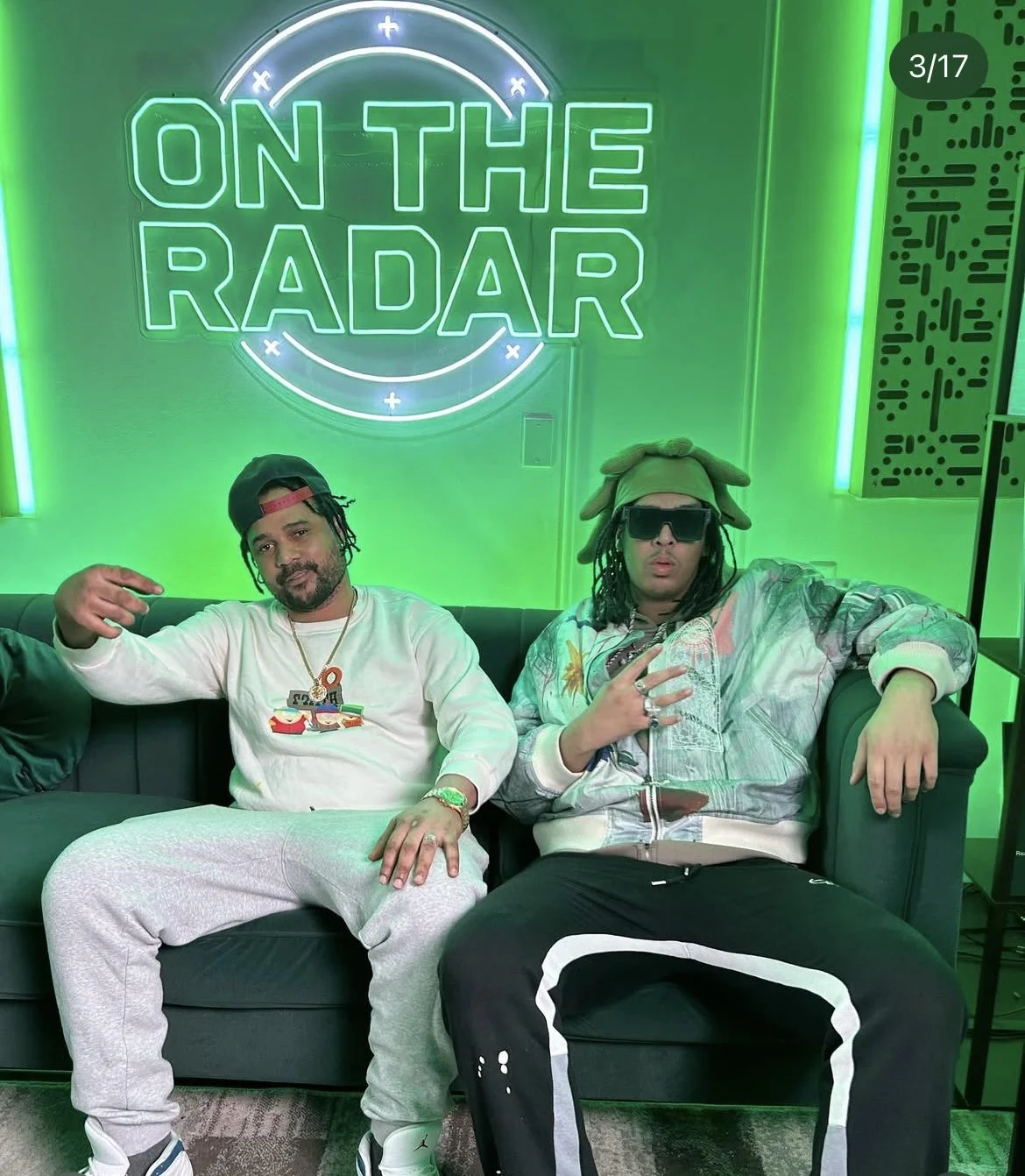 Vicer and another man sitting on a green couch in front of a neon sign that says 'On The Radar.' One man is wearing a white sweatshirt, gray sweatpants, and a black cap backward.