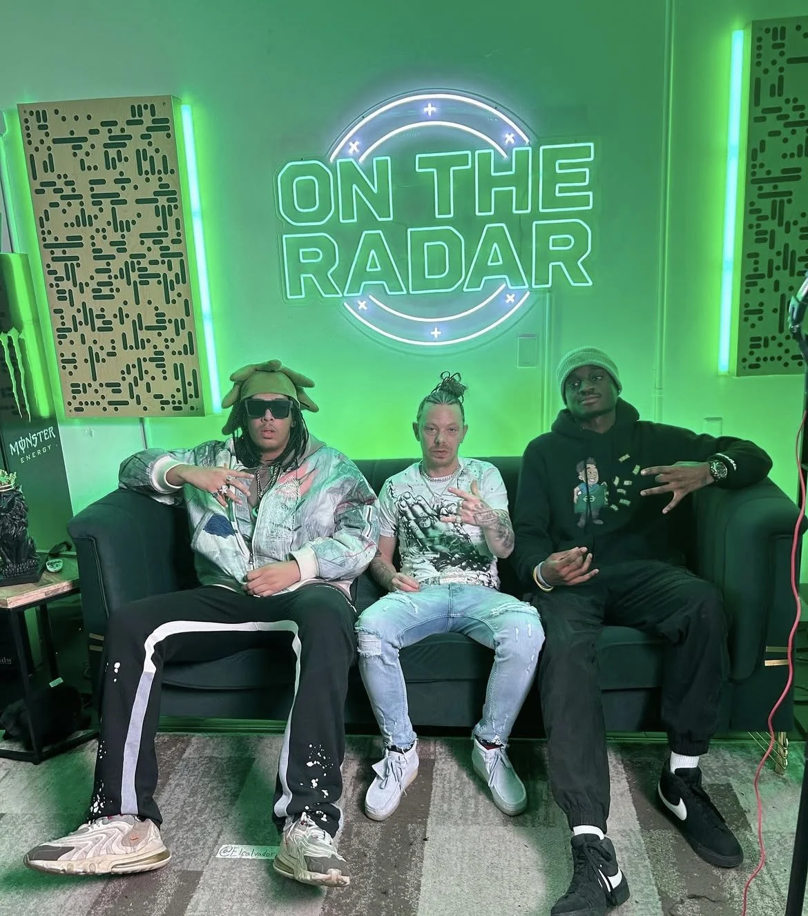 Vicer and two other men sitting on a black couch in front of a bright green neon sign that reads 'On The Radar' with a circular design. Vicer wears sunglasses, a beige hat, a shiny patterned jacket, and black pants with white stripes.