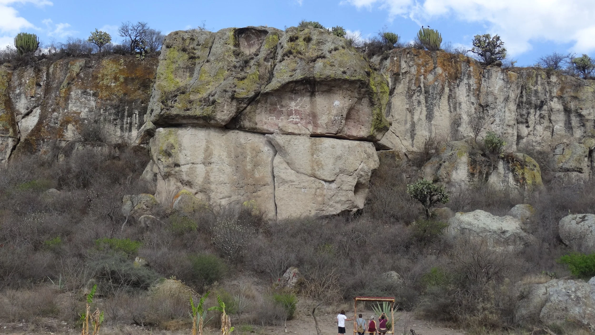 Group of four people sitting on a bench and taking a photo of a large rocky cliff with sparse vegetation and cacti.