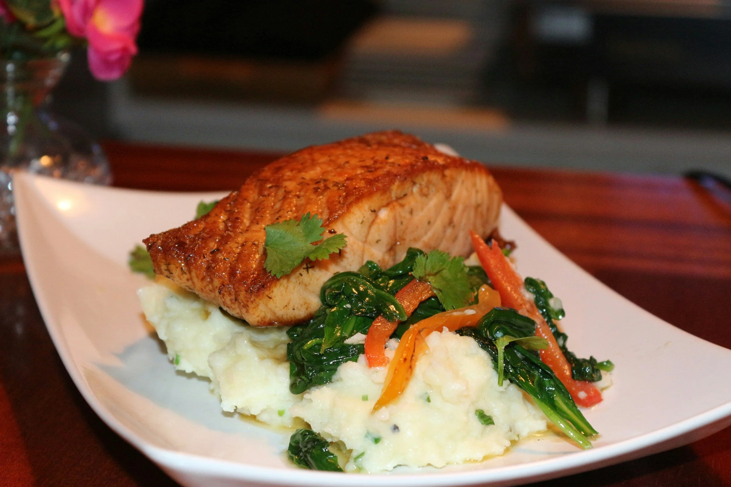 A plate with a cooked piece of salmon, mashed potatoes, and sautéed greens with carrots, garnished with cilantro, on a white rectangular dish.