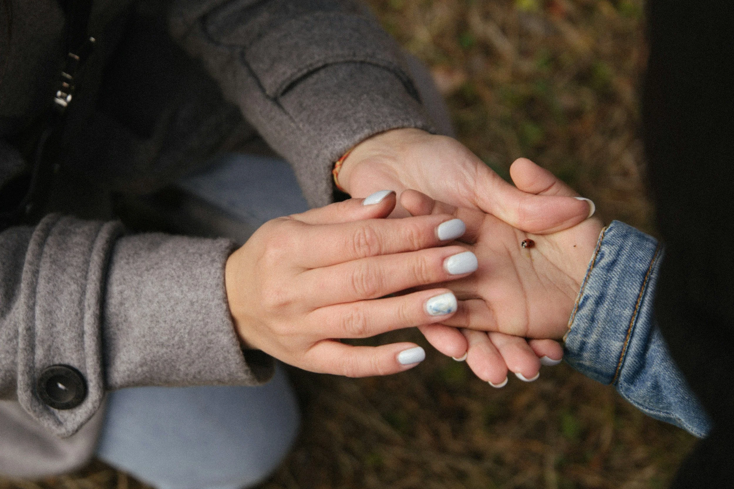 Two people holding hands, one with neatly manicured nails and the other with a small ring and a bracelet, outside on a natural ground surface.